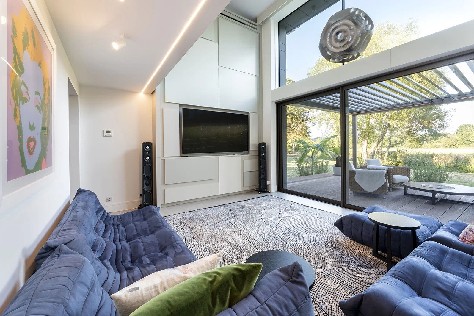 Modern living room with blue couches, a large flat-screen TV, tall speakers, a patterned rug, and large glass doors leading to an outdoor patio with outdoor seating, greenery, and trees.