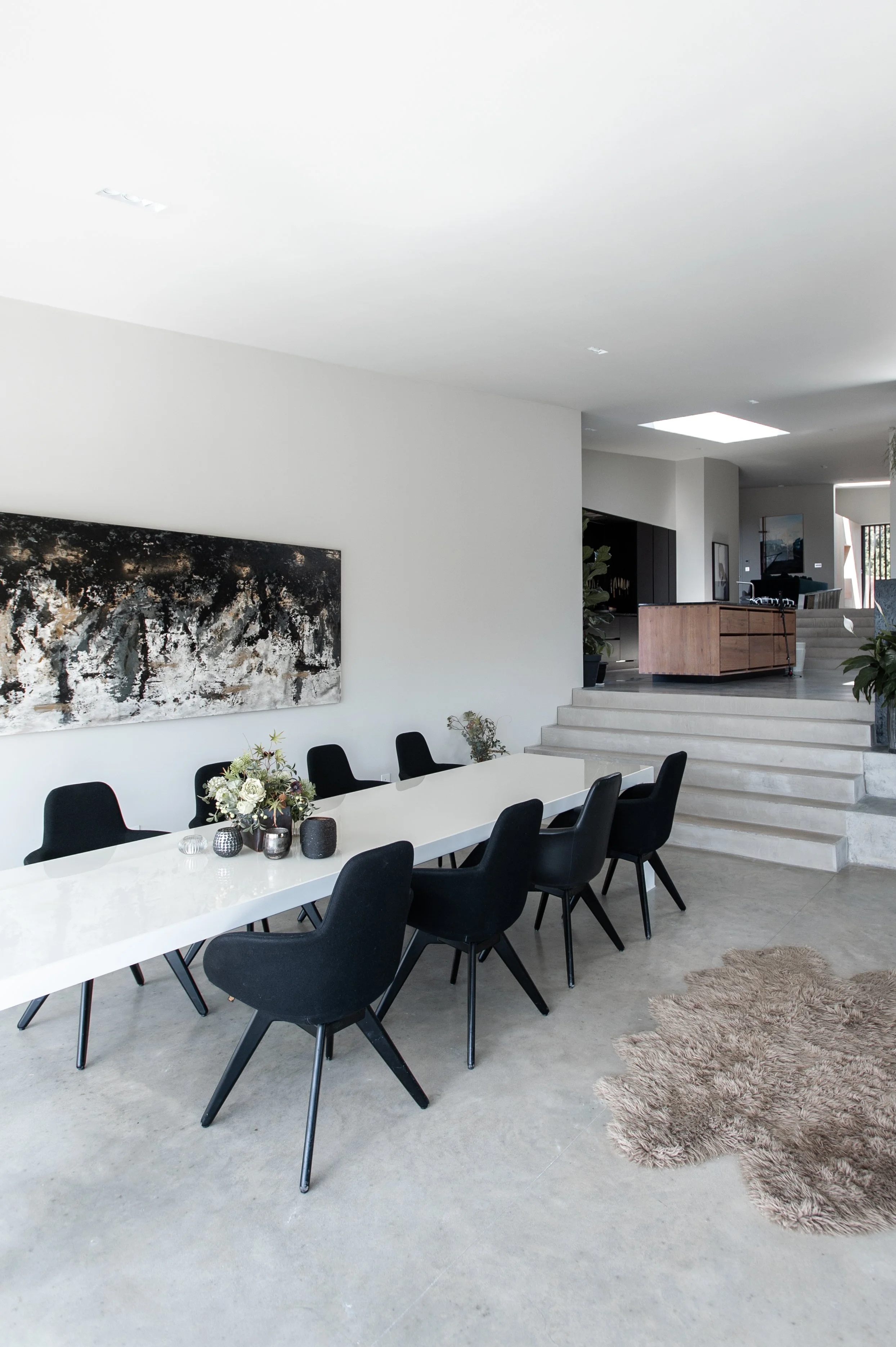 Modern dining room with a white table, black chairs, floral centerpiece, abstract wall art, and stairs leading to an open kitchen area.