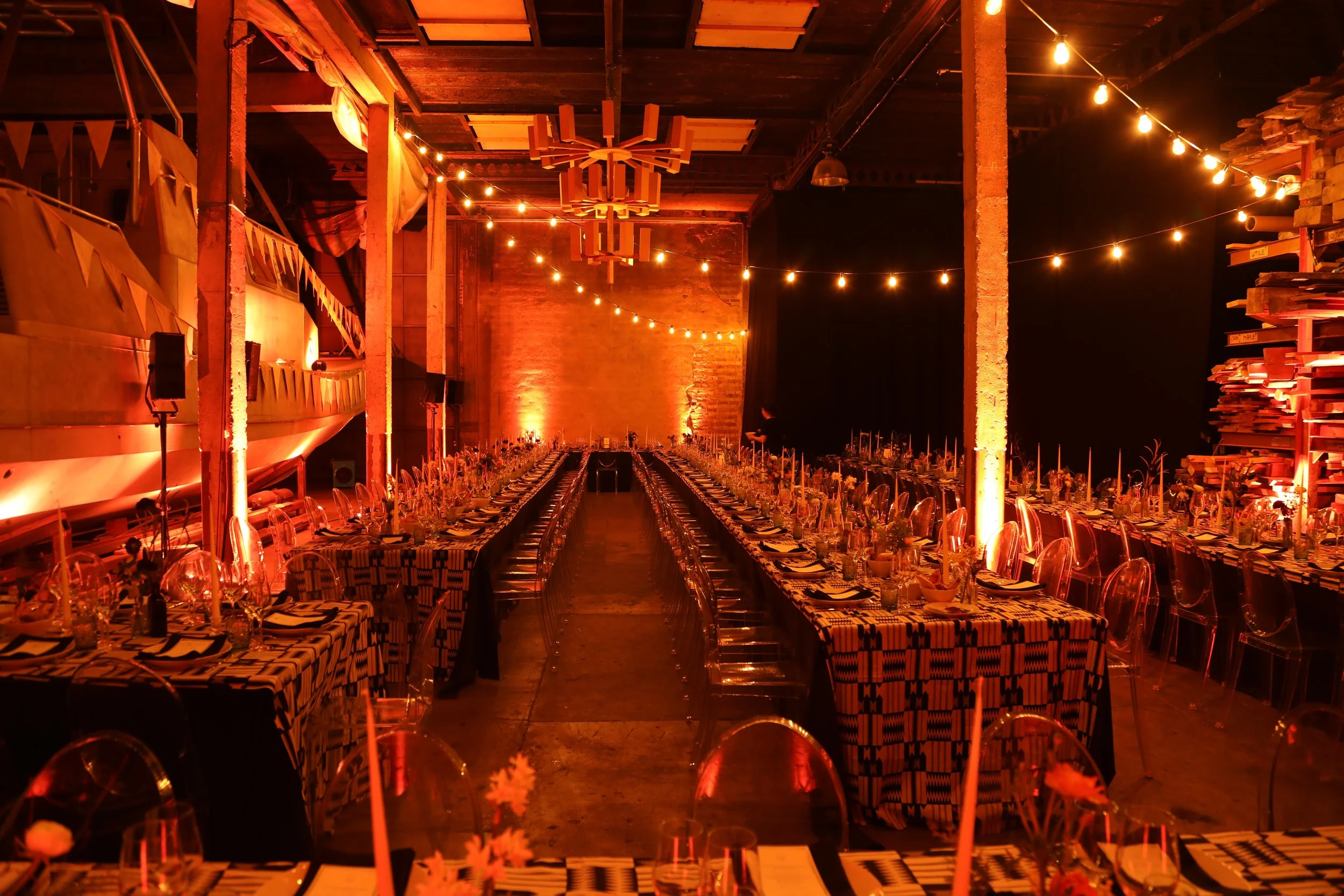 Empty banquet hall decorated for a celebration with long tables covered in black and white patterned tablecloths, clear plastic chairs, string lights hanging from the ceiling, and a wooden wall and bookshelves on the right side, illuminated by warm o