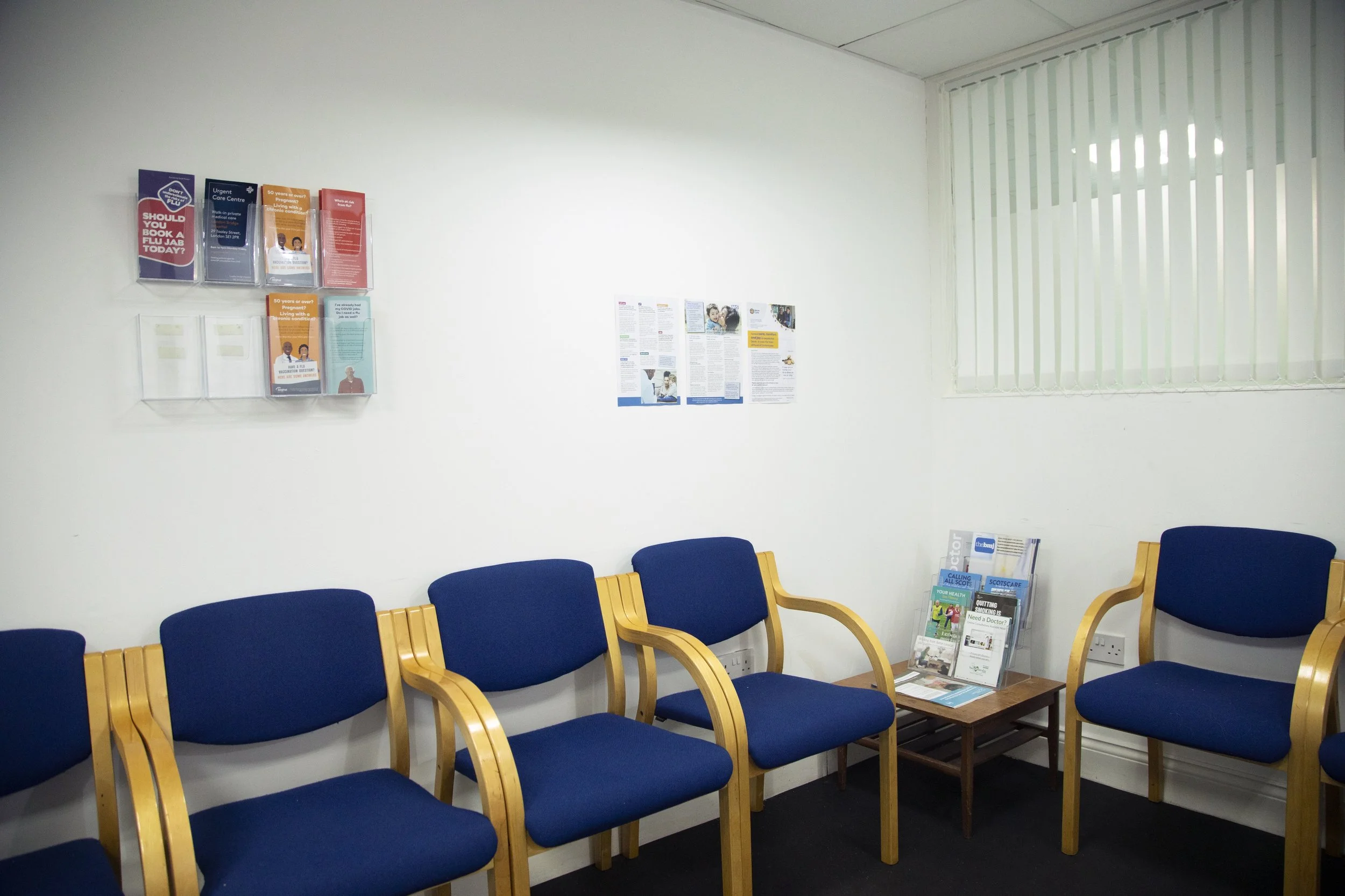 Waiting room with five blue chairs with wooden armrests and a small table holding magazines, white wall with brochures and posters, window with vertical blinds.