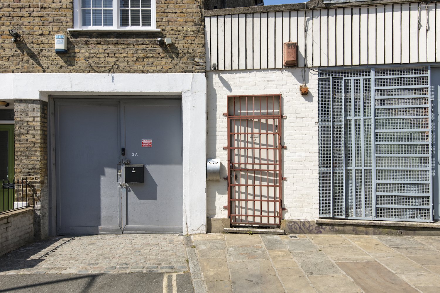 Facade of a building with a gray metal garage door, a metal security gate, and a partially painted brick wall, with a window above and a sidewalk in front.