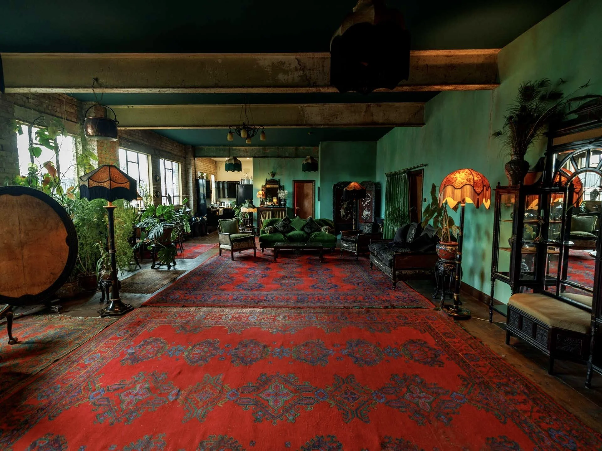 Interior of a vintage-style living room with green walls, large red patterned rugs, antique furniture, floor lamps, and potted plants, illuminated by natural light from multi-paned windows.