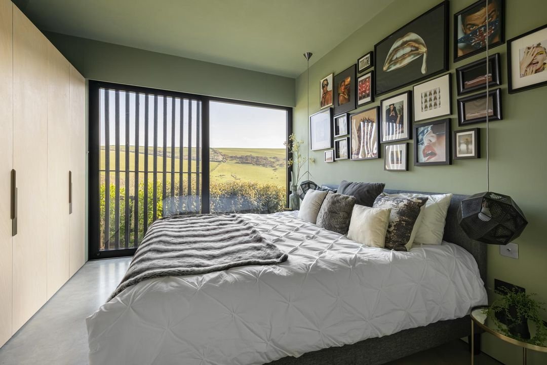 Modern bedroom with green walls, gallery wall with framed art above a bed, large window with vertical blinds showing a green landscape, off-white wardrobe, and a small side table with a plant.