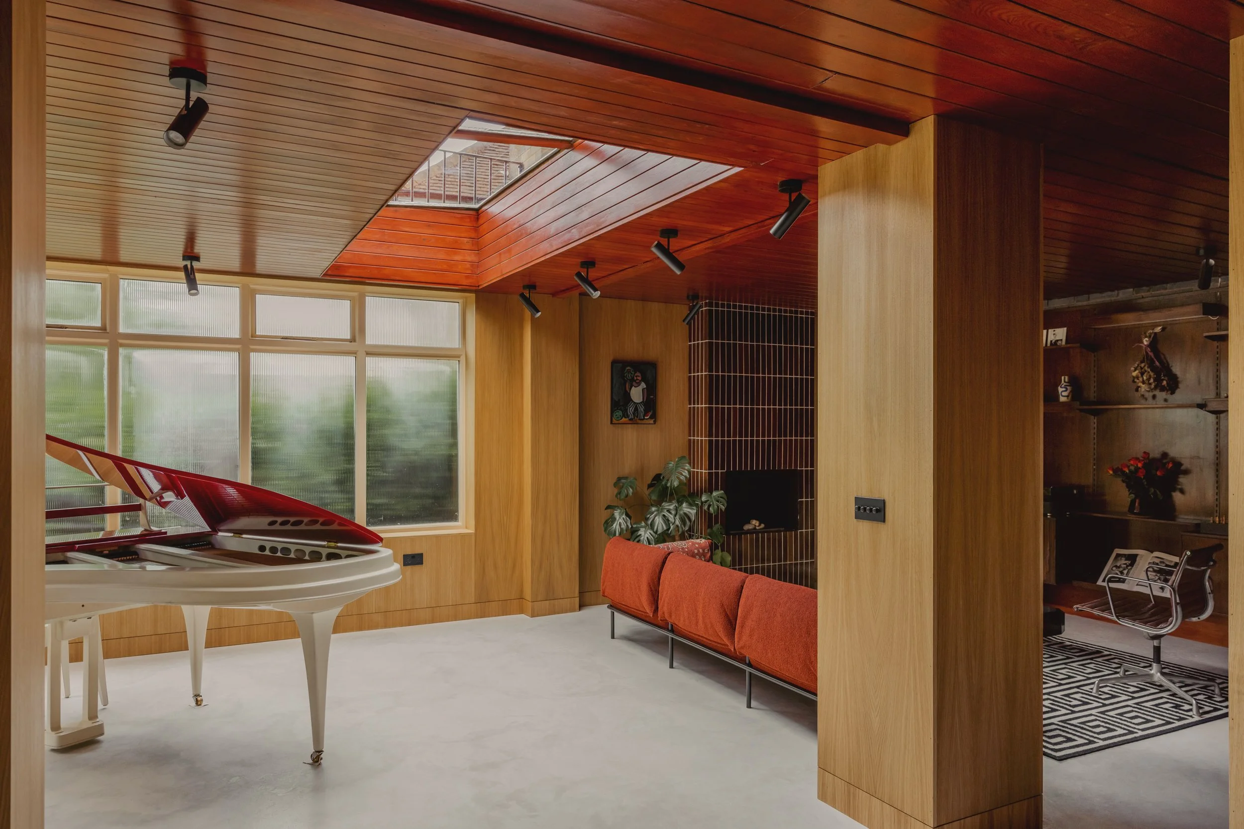 Modern living room with a white grand piano, large windows, wooden walls and ceiling, ceiling spotlights, a red sofa, art on the wall, indoor plants, and a small sitting area with a chair and bookshelf.