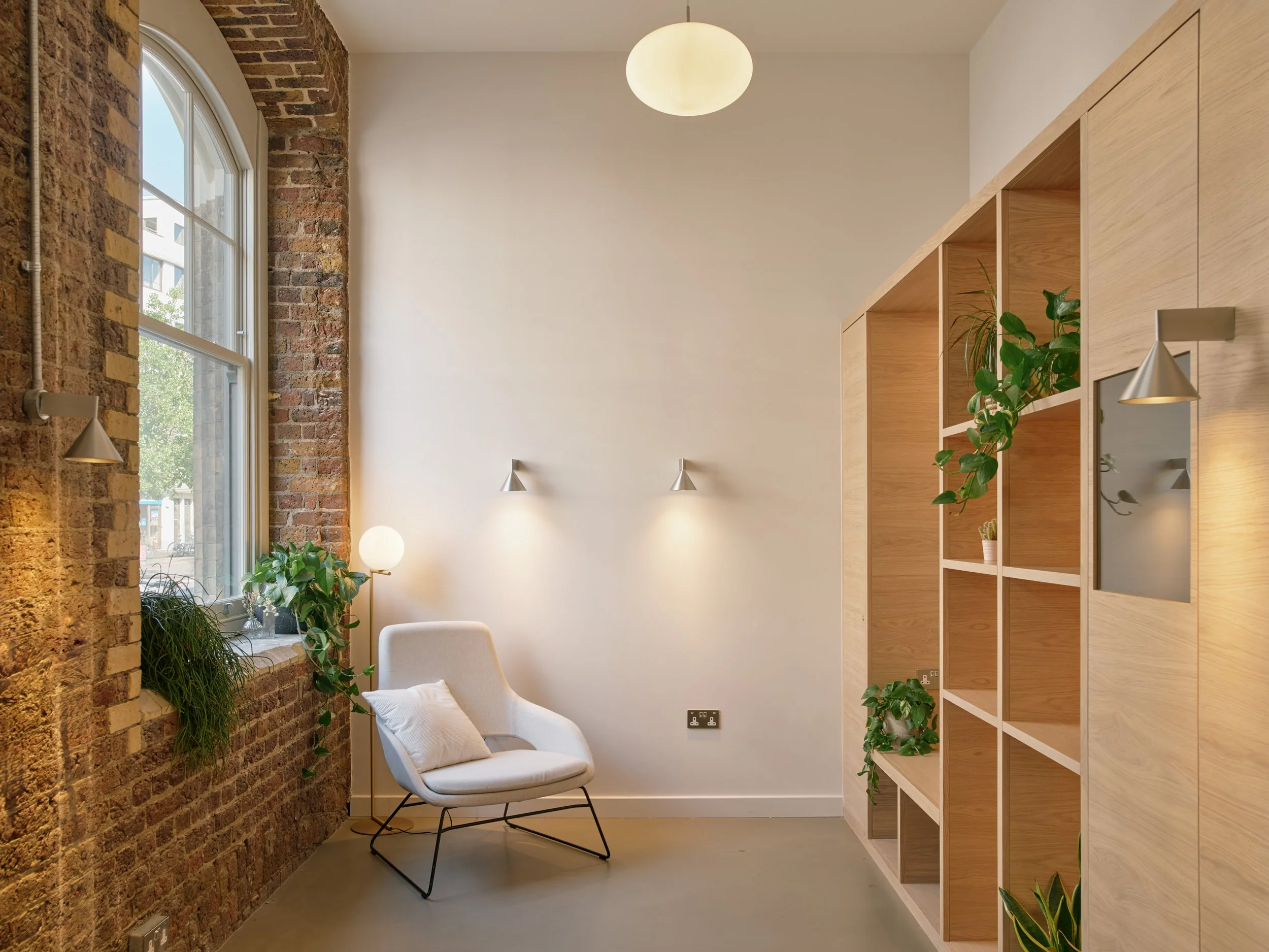 A cozy interior corner with a large brick wall and a window with green plants on the windowsill. There is a white modern armchair with a cushion, a wall-mounted light fixture, a ceiling light, and a wooden shelving unit with plants and decor.
