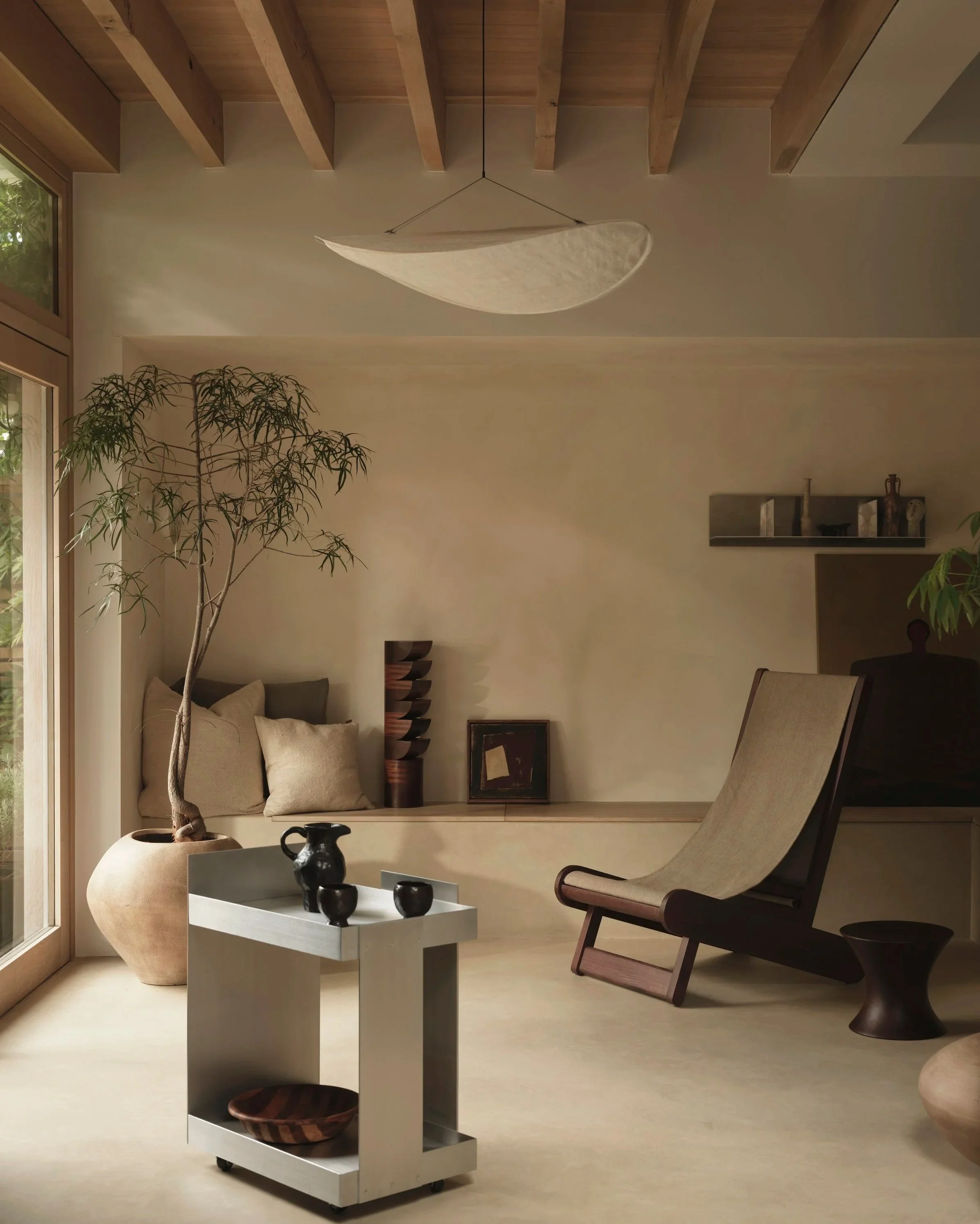 Minimalist living room with beige walls, wooden ceiling beams, a large potted tree near the window, a modern metal cart with black pottery, a beige lounge chair, and decorative objects on the wall shelf.