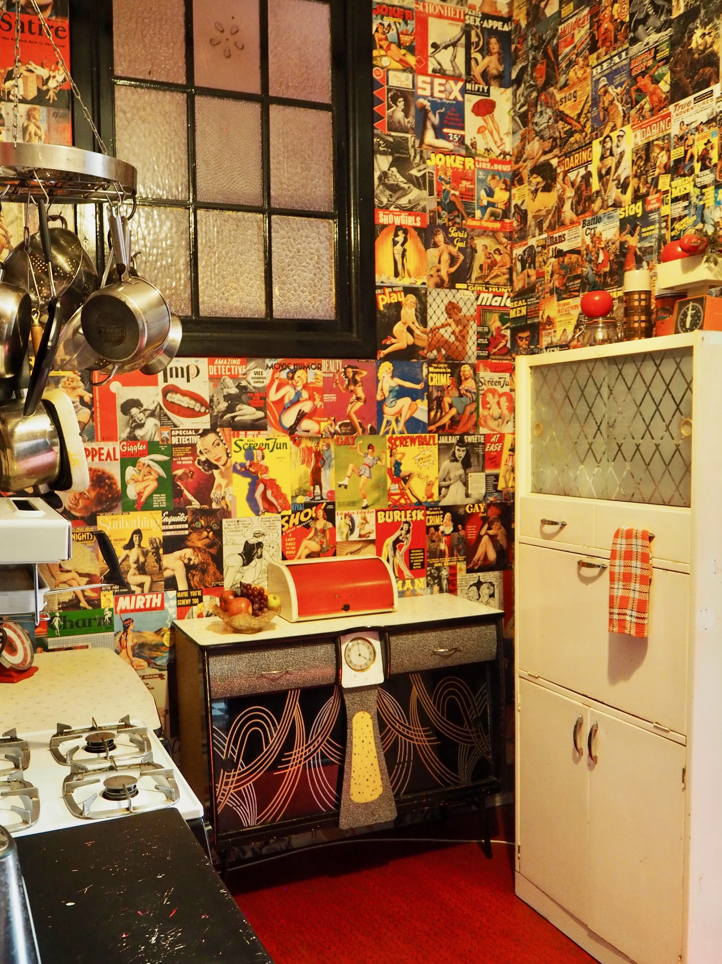 A vintage-style kitchen corner featuring a wall covered with colorful vintage comic and pin-up magazine posters, a white refrigerator with a red and white checkered towel hanging on the door, a small black and white oven with a clock, and a fruit bow
