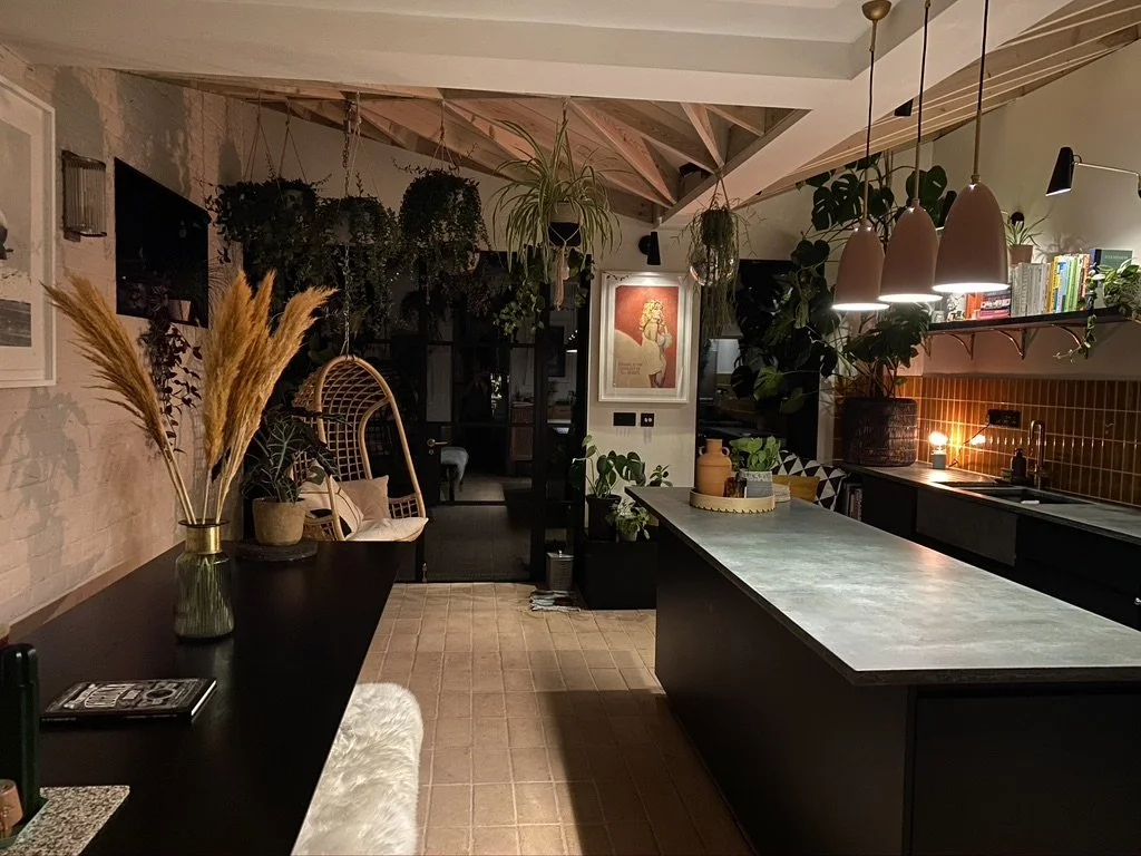 A cozy kitchen with a large island, pendant lights, shelves with books, numerous plants hanging and on surfaces, a framed artwork on the wall, and dim warm lighting.