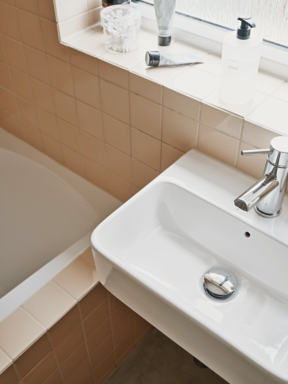 A white bathroom sink with a chrome faucet, with pinkish-brown tiled walls and windowsill with toiletries, including lotion, toothpaste, and cream.