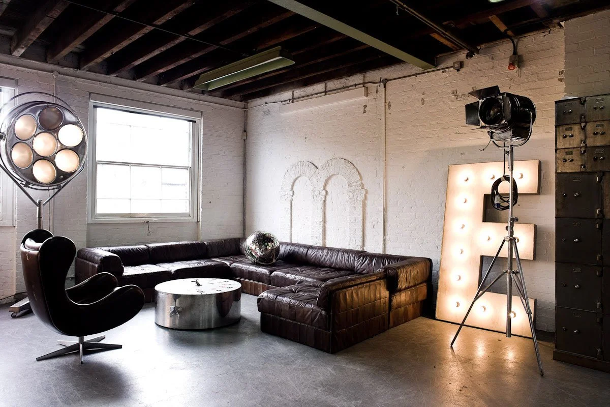Industrial-style living room with a large L-shaped leather sofa, a black modern armchair, a metallic round coffee table, studio lighting, a large illuminated letter 'E', and a wall of lockers.