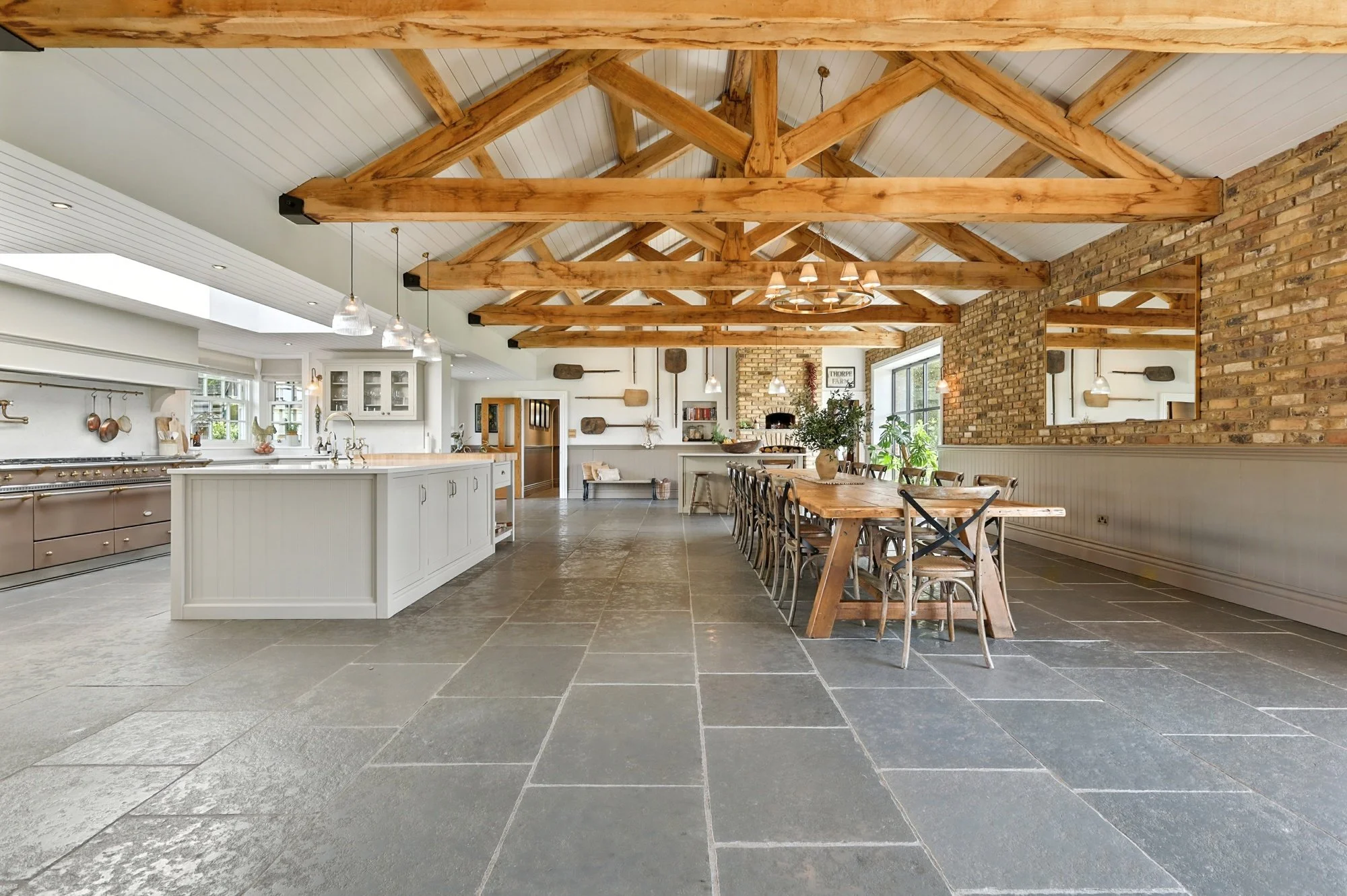 Spacious kitchen and dining area with exposed wooden beams, brick wall, large window, white cabinets, and a long wooden dining table surrounded by chairs.