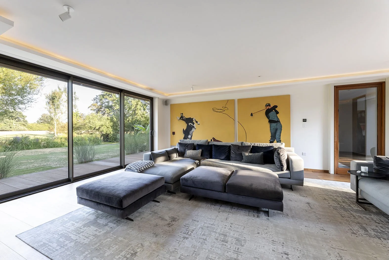 Modern living room with large glass sliding doors, a gray sectional sofa, black and gray throw pillows, a patterned blanket, and a rug. Art depicting a golfer and a woman on a yellow background hangs on the wall.