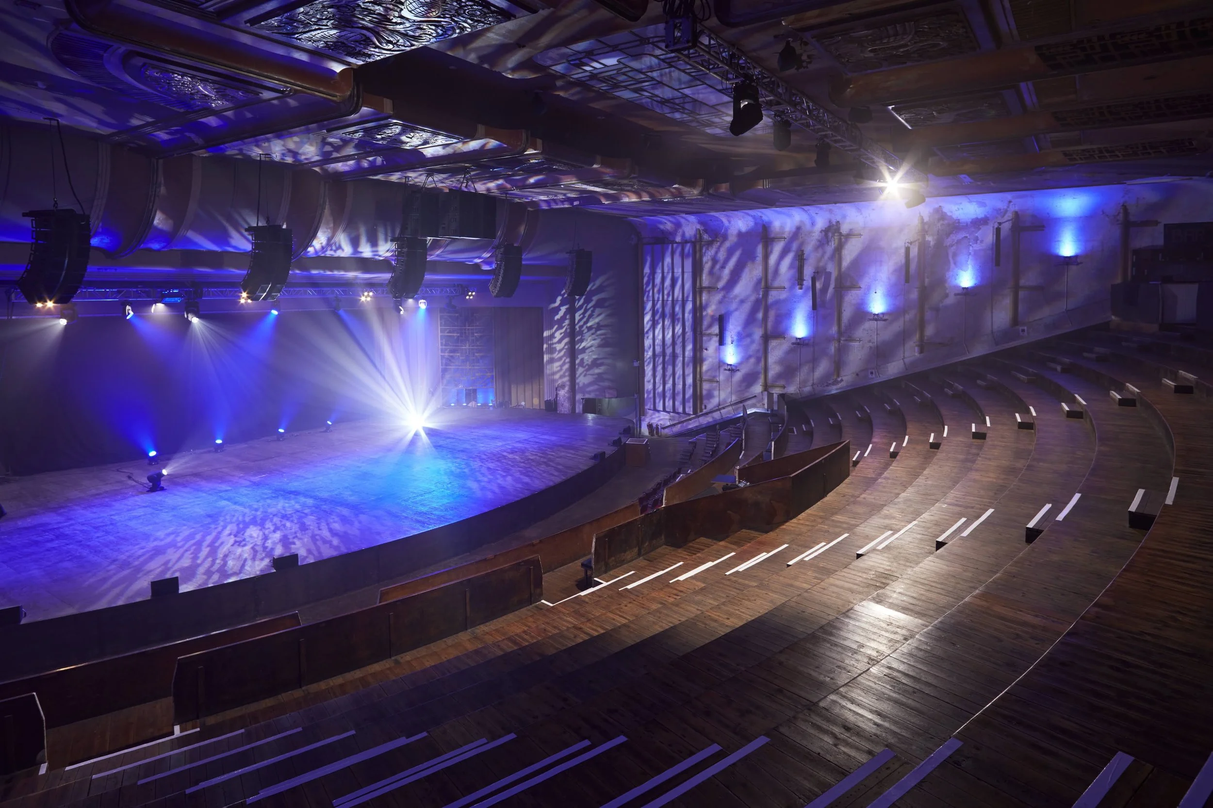 Empty auditorium with illuminated stage and wooden seating area, blue and purple lighting, and decorative wall lights.