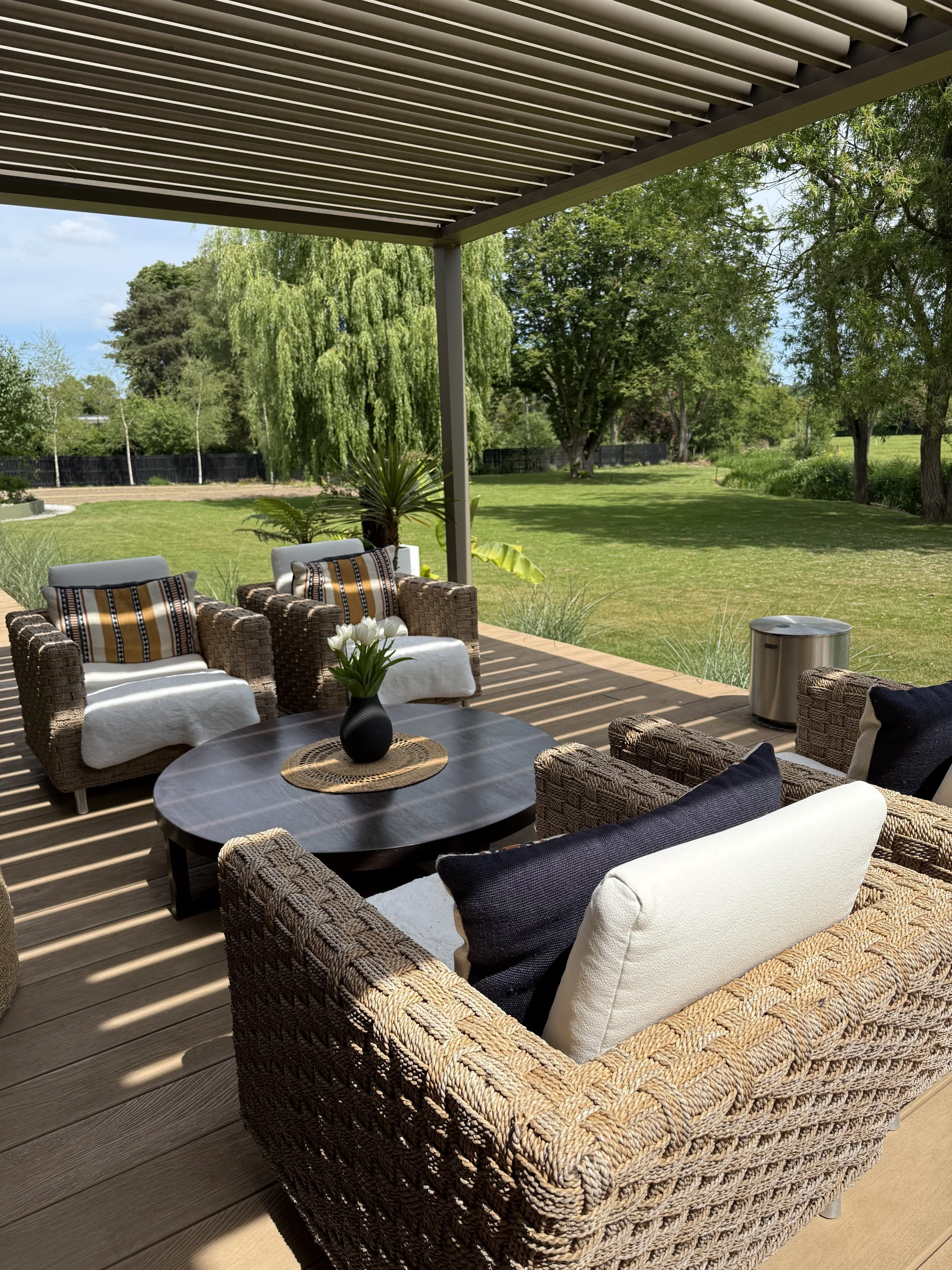 Outdoor patio area with wicker and cushioned chairs arranged around a round table, a vase of white flowers, and a green lawn with trees in the background.