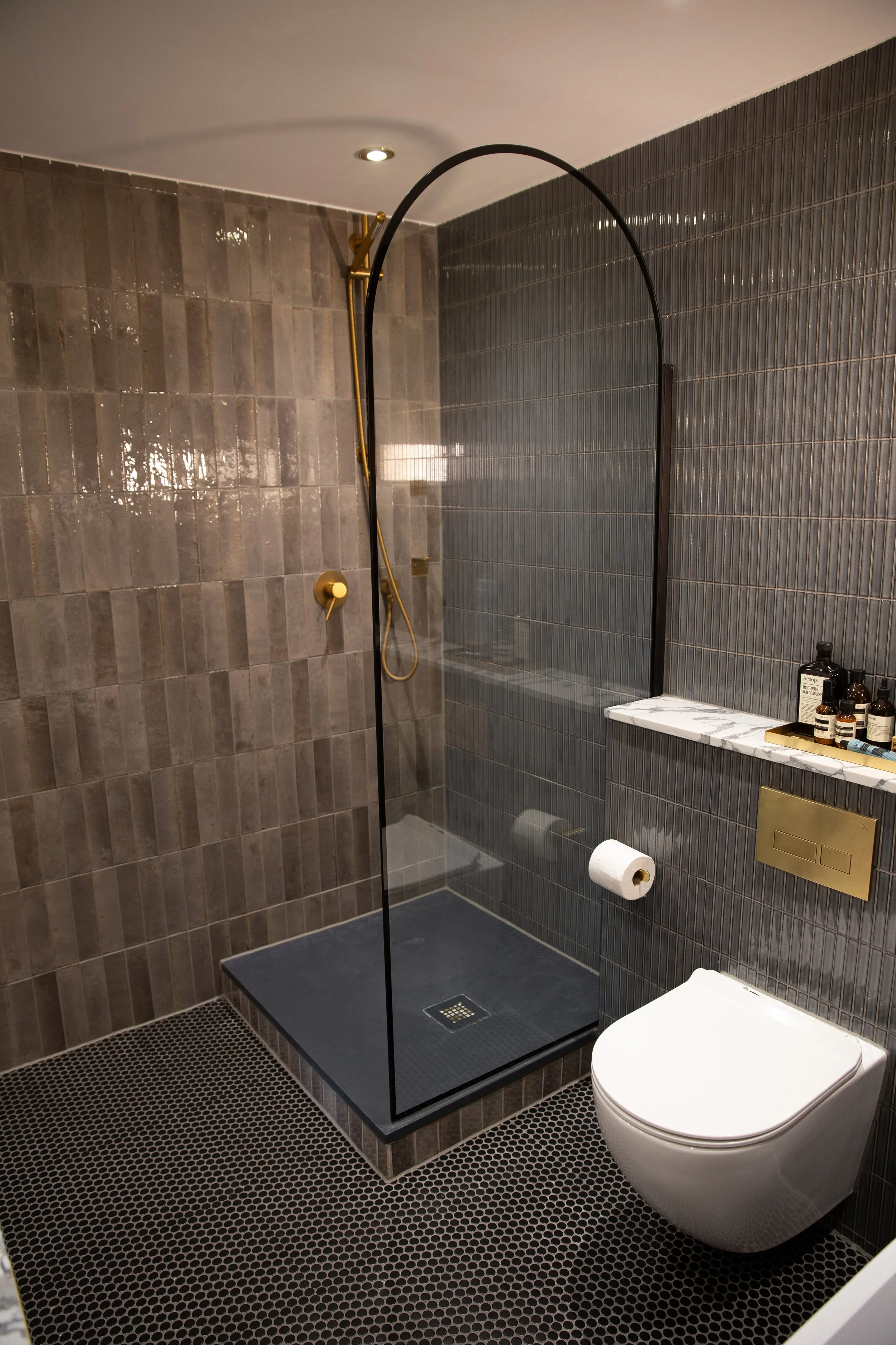 Modern bathroom with walk-in shower featuring a black glass partition, dark gray tiled walls, and a white wall-mounted toilet. The shower has a brass shower head and hose, and bathroom essentials are on a small shelf with marble details. The floor ha