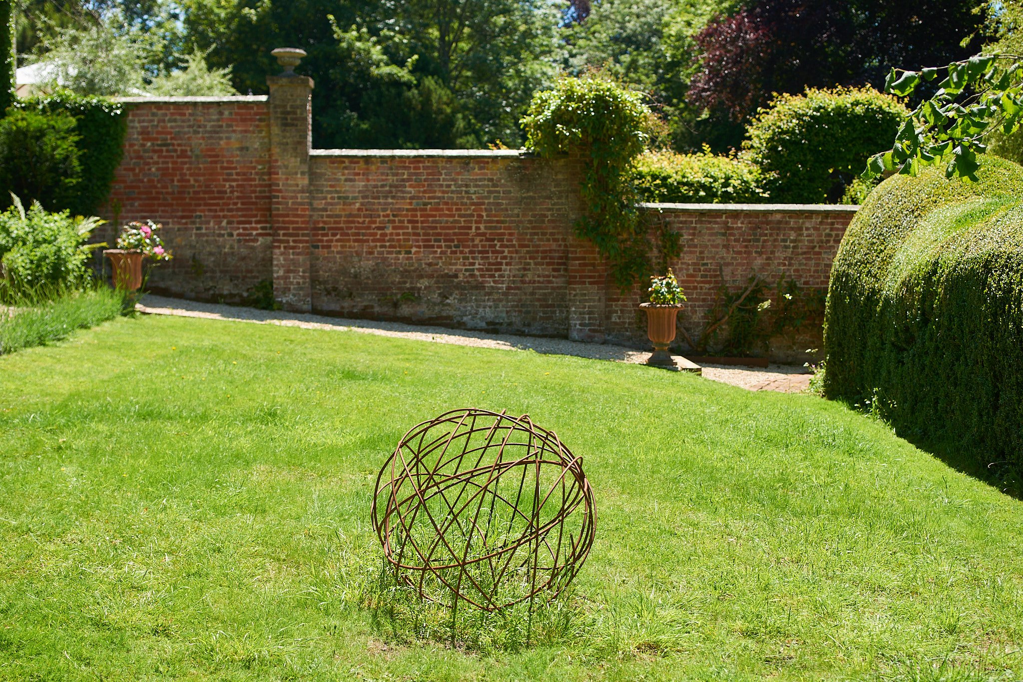 A well-maintained garden with green grass, potted plants, and a red brick wall in the background. There is a metal decorative sphere in the center of the lawn.