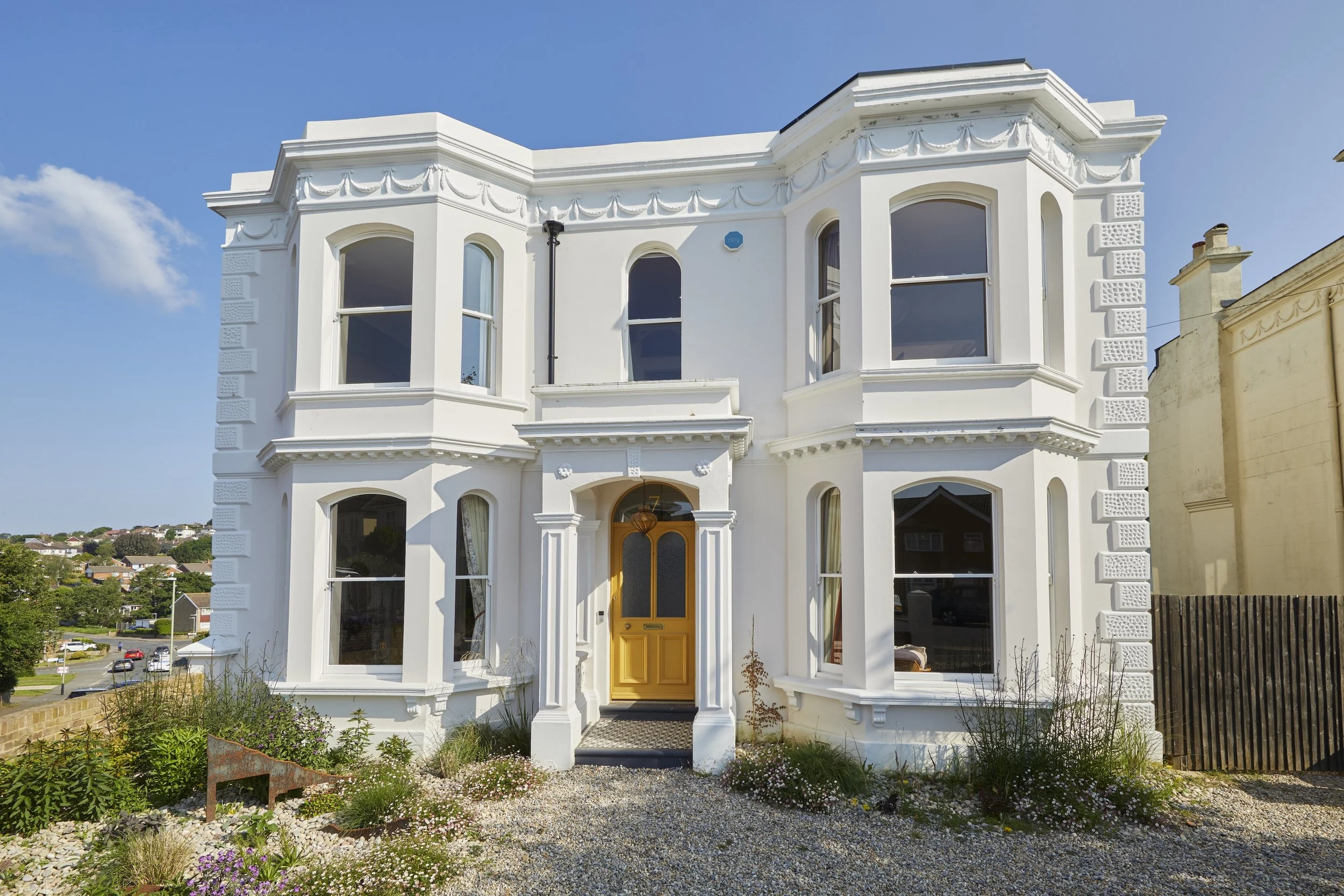 A white Victorian-style two-story house with decorative trim, large windows, and a yellow front door, surrounded by a gravel yard and garden with flowers and plants.