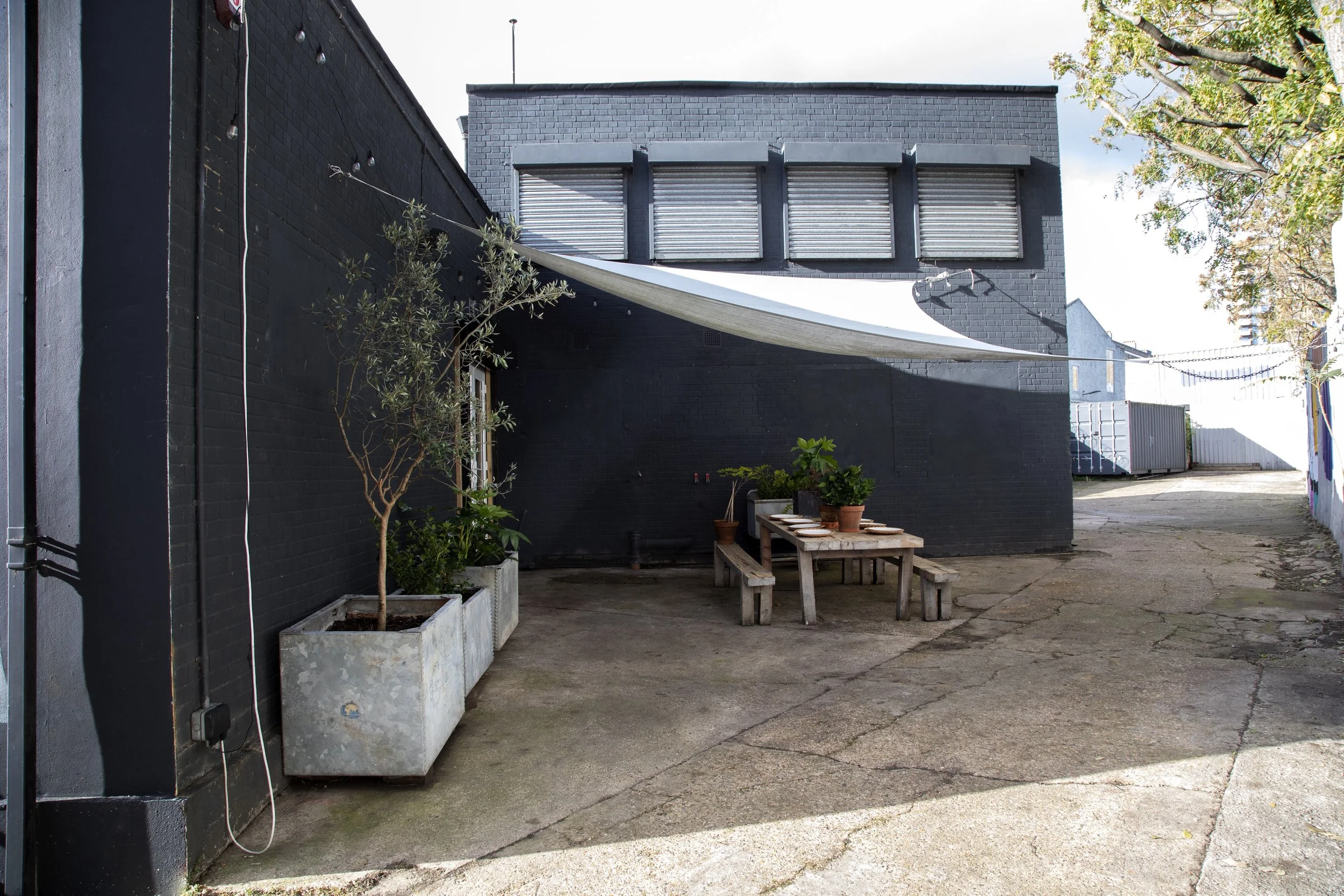 An outdoor alleyway scene with a black brick building in the background, featuring three small windows with blinds, a white shade sail canopy, a wooden picnic table with benches, potted plants on the table, and a large pot with a small tree on the le