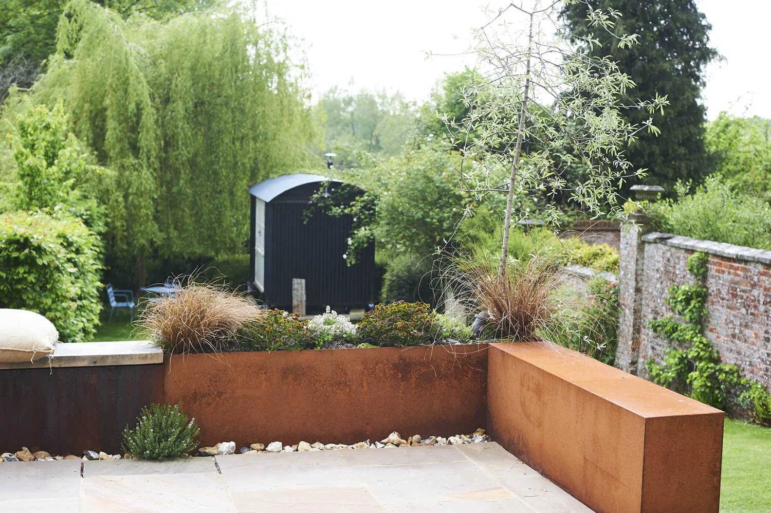 View of a backyard garden with a patio, plants, trees, a black shed, and a brick wall.