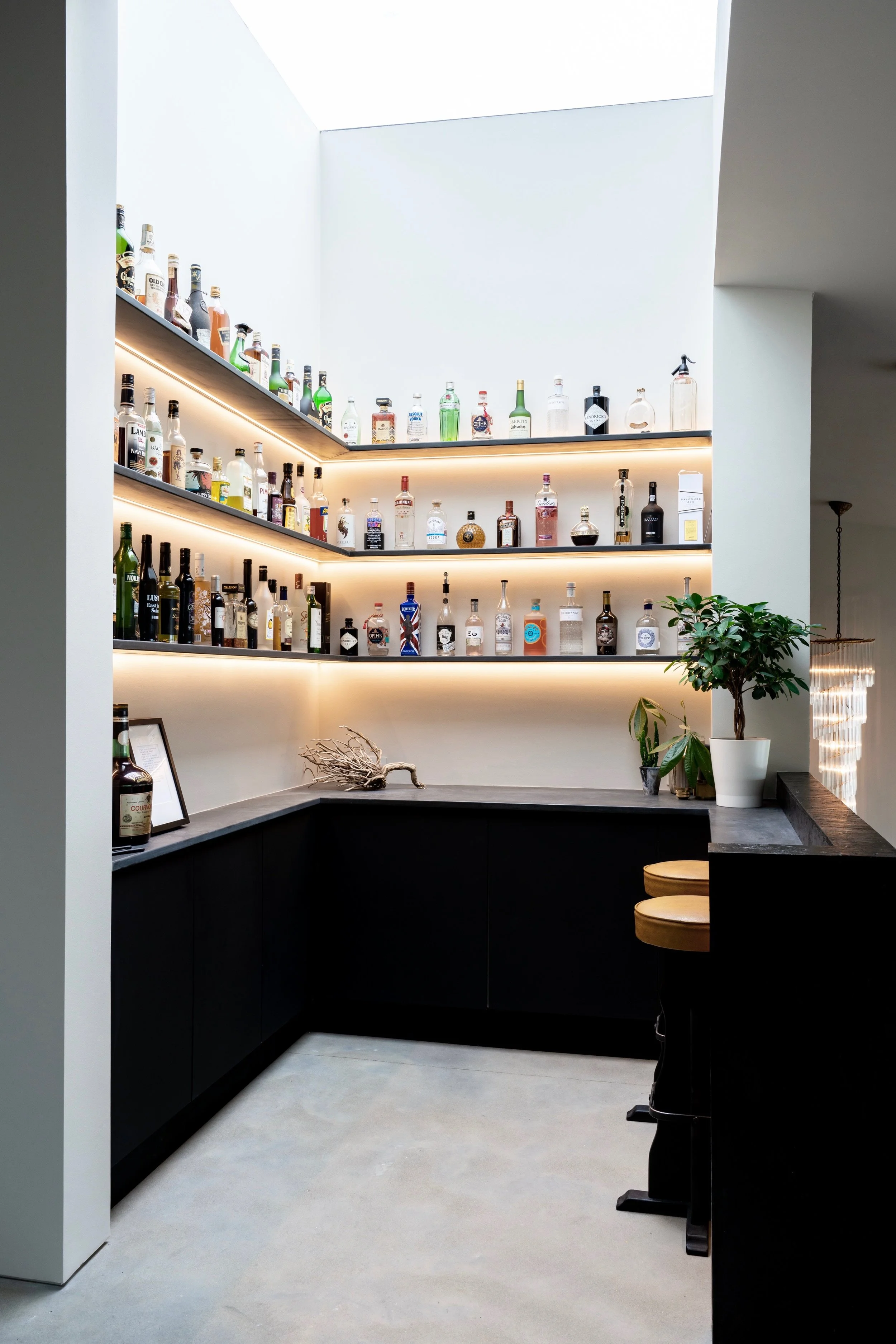 A modern home bar with black cabinetry, a light-colored countertop, and three wooden bar stools. Shelves with backlit lighting display a variety of liquor bottles. A potted plant, a framed picture, and decorative branches sit on the counter.