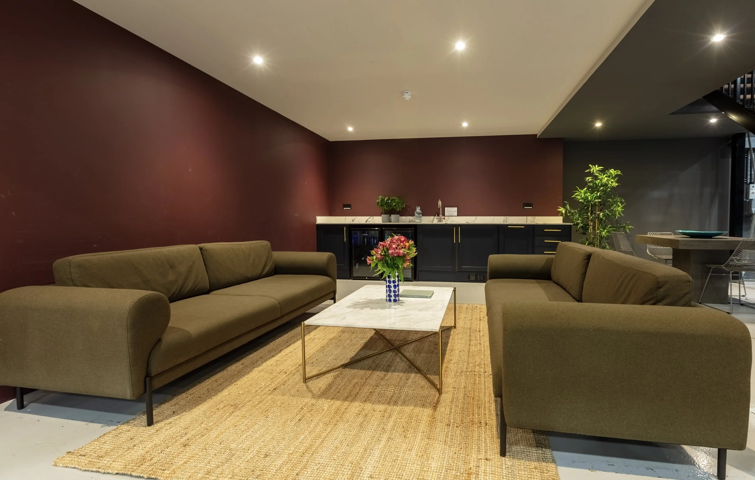 Living room with two olive green sofas, a marble coffee table with a blue and white vase holding pink flowers, a built-in bar with a dark cabinet, countertop, and plants in the background.