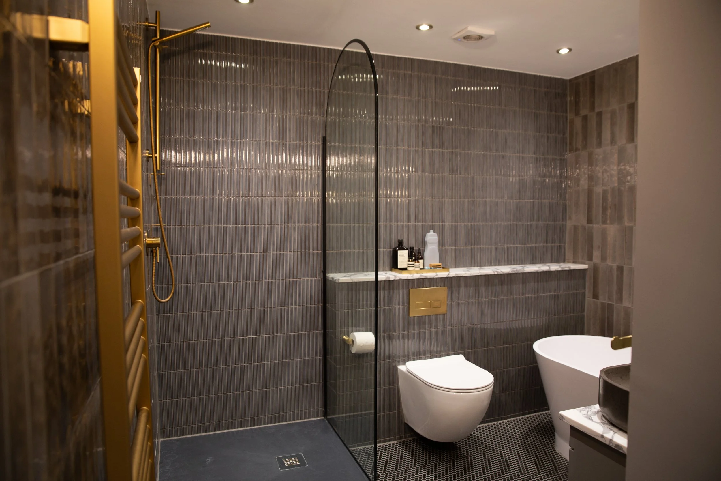 Modern bathroom with dark gray tiled walls, a wall-mounted toilet, a freestanding bathtub, and a marble shelf with toiletries. Gold accents include a heated towel rail and flush plate. Recessed ceiling lights illuminate the space.