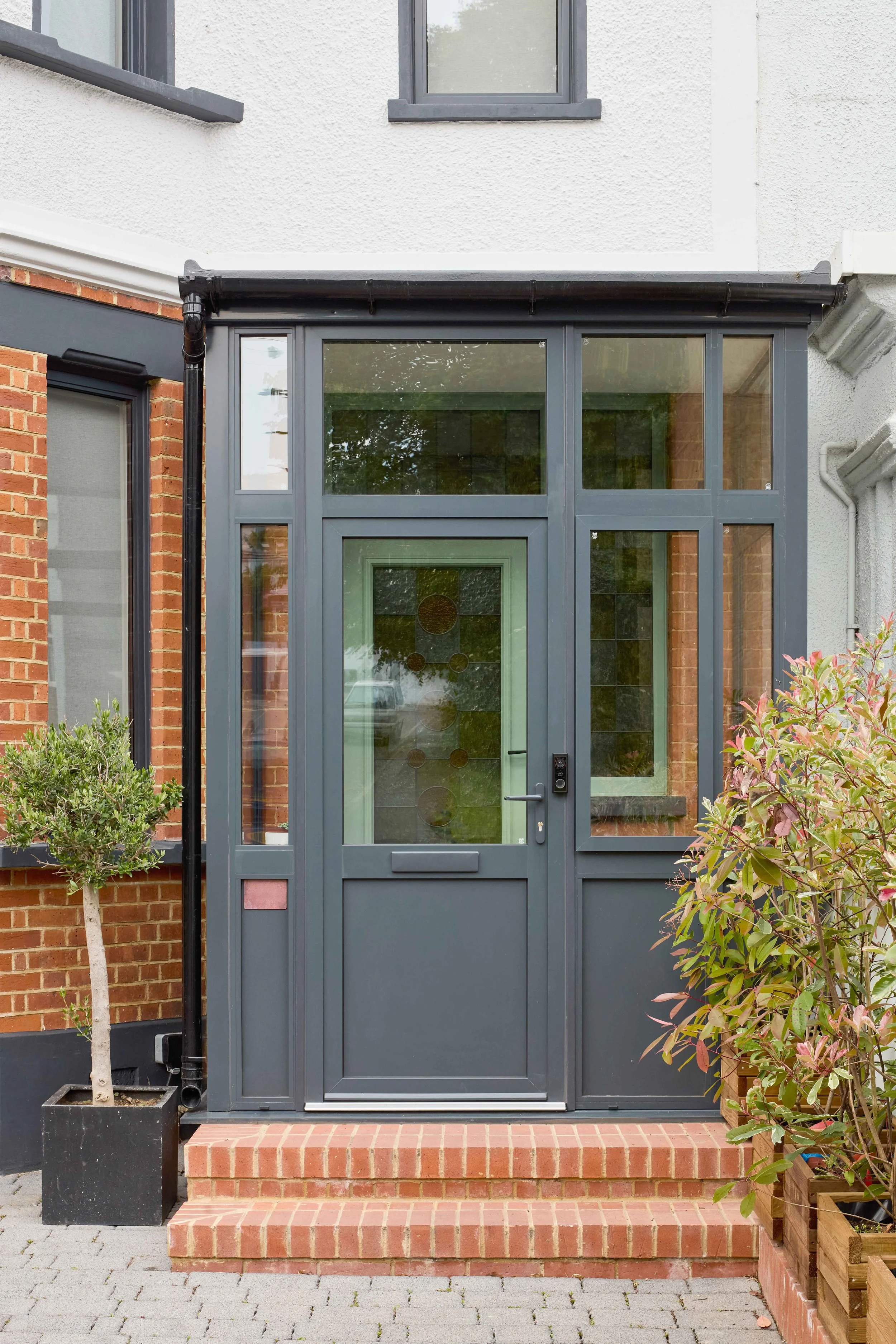 A modern enclosed porch with gray metal framing, brick steps, and a glass door; flanked by potted shrubbery and with windows on the upper level.