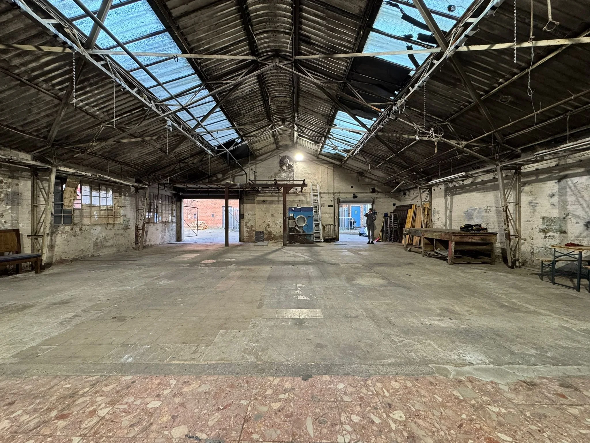 An empty industrial warehouse with a high, metal arched ceiling and large open entrance doors, showing a person inside.
