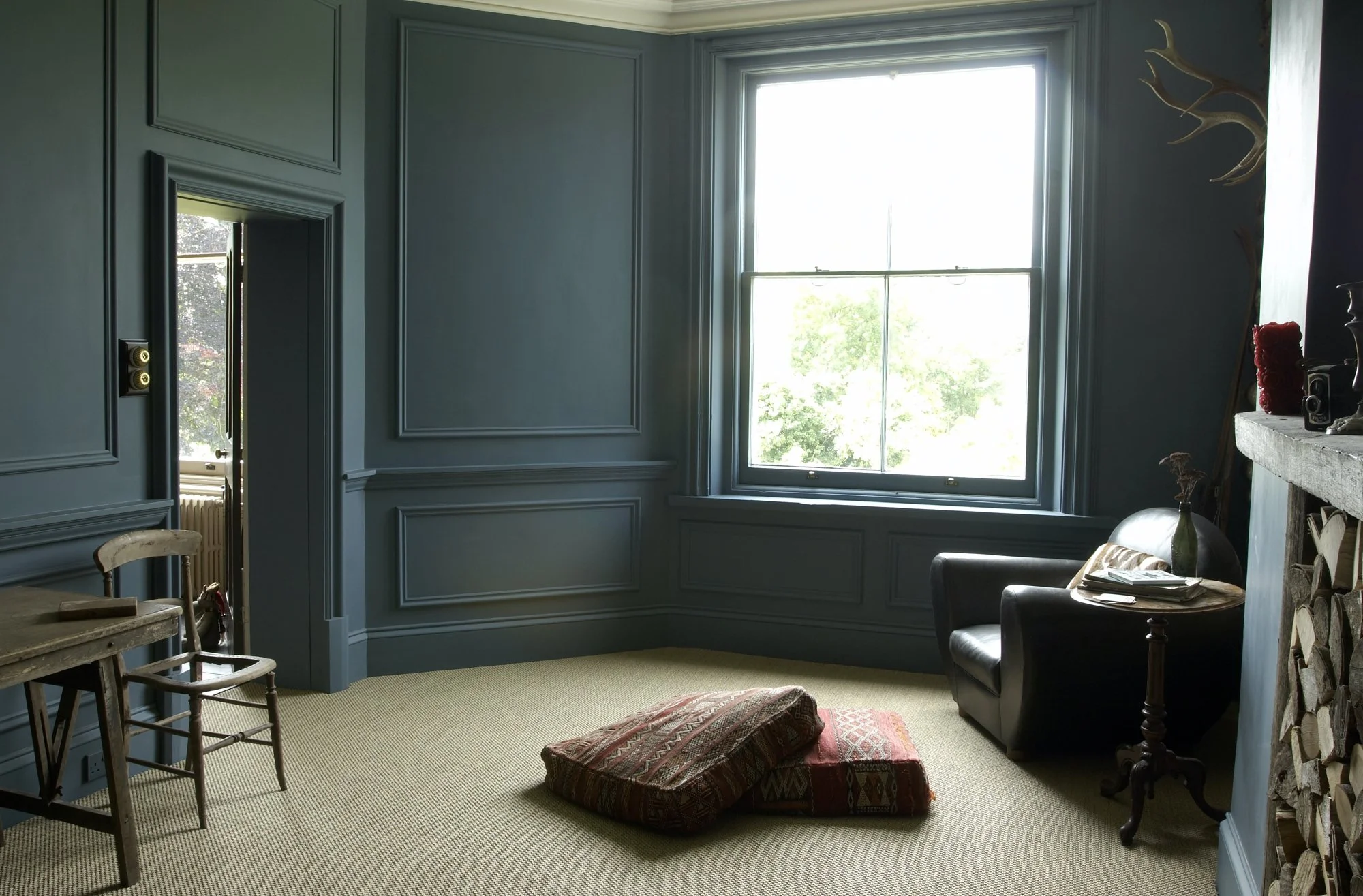 Room with blue paneled walls, a large window with greenery outside, a black armchair with a side table holding books and a vase, a small desk with a chair, and a pile of decorative cushions on the beige carpeted floor.