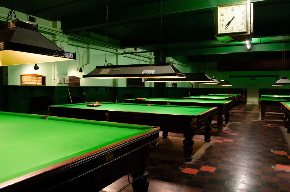 Multiple snooker tables with green cloths in an indoor billiards hall, illuminated by hanging lights, with vintage wall clock visible at the top.