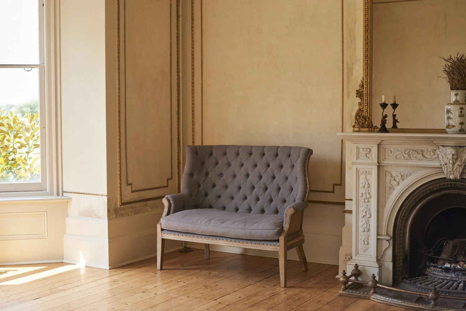 A vintage-style interior room with a tufted light gray armchair, ornate cream-colored fireplace with black interior, candle holders, and a potted plant on top; bright sunlight coming through a window; wooden floor and decorative wall molding.