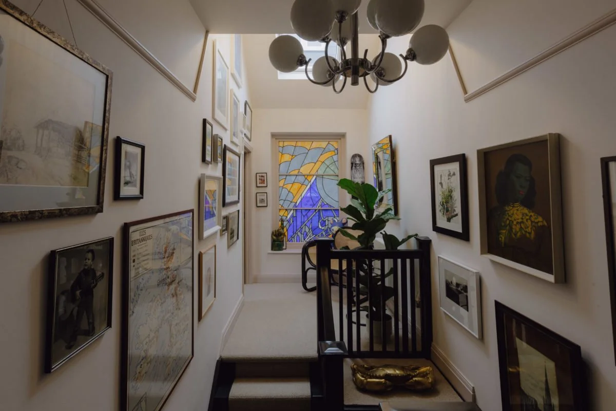Indoor hallway decorated with framed pictures, a chandelier hanging from the ceiling, a stained glass window at the end, a potted plant, and a chair with a cushion.