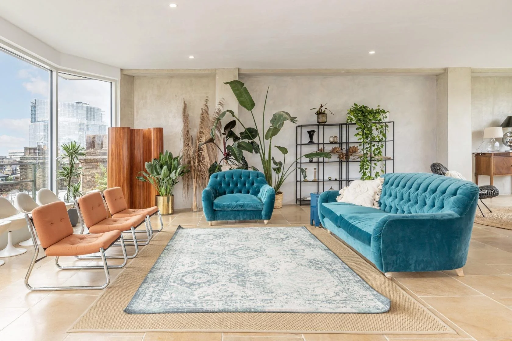 Bright living room with large windows, blue tufted sofa, matching armchair, beige chairs, greenery, and decorative shelving.