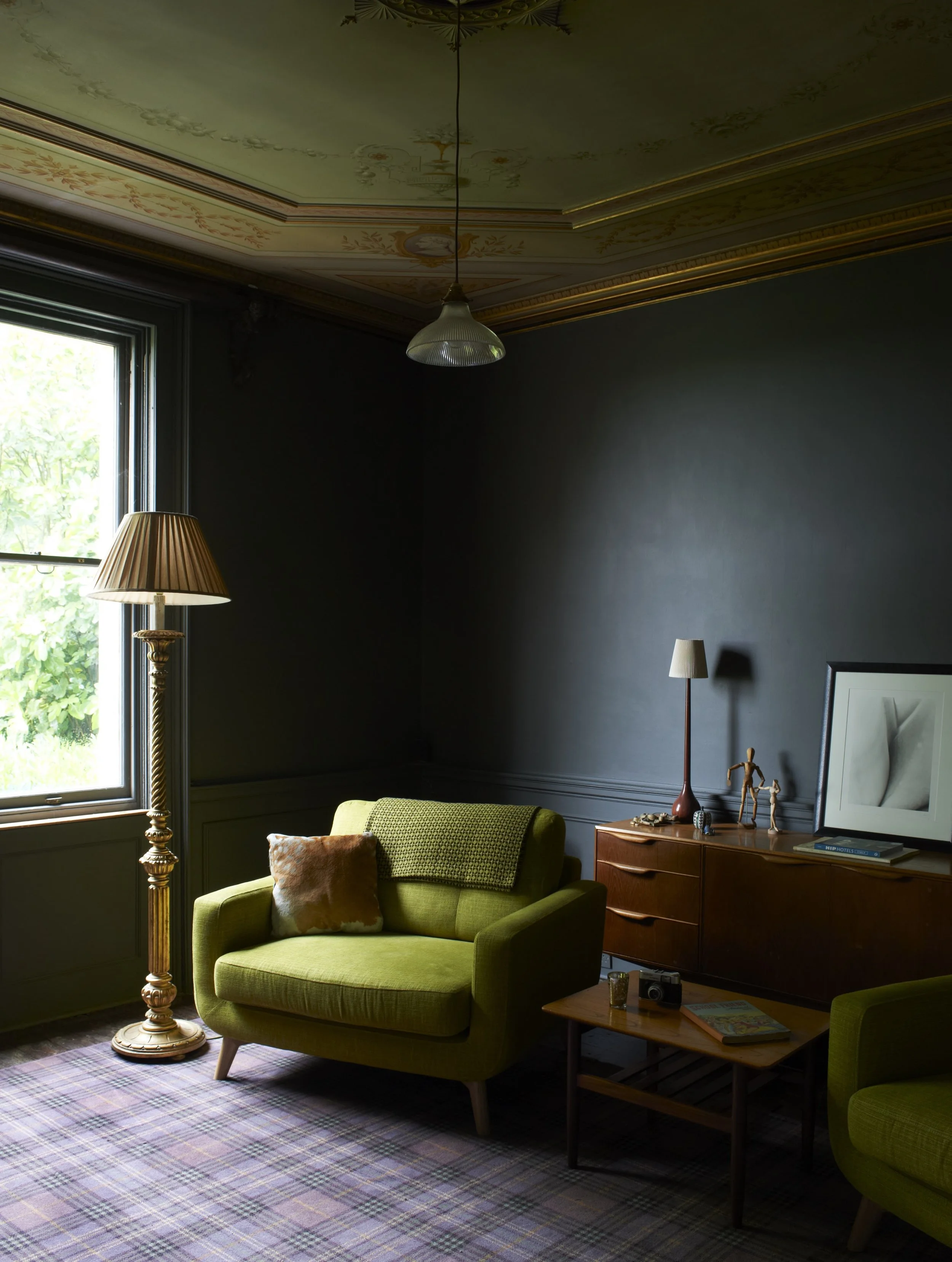 A vintage-style living room with dark walls, green armchairs, a tall floor lamp, and a wooden sideboard with decorative objects. A window lets in natural light, revealing a plaid carpet and artwork on the sideboard.