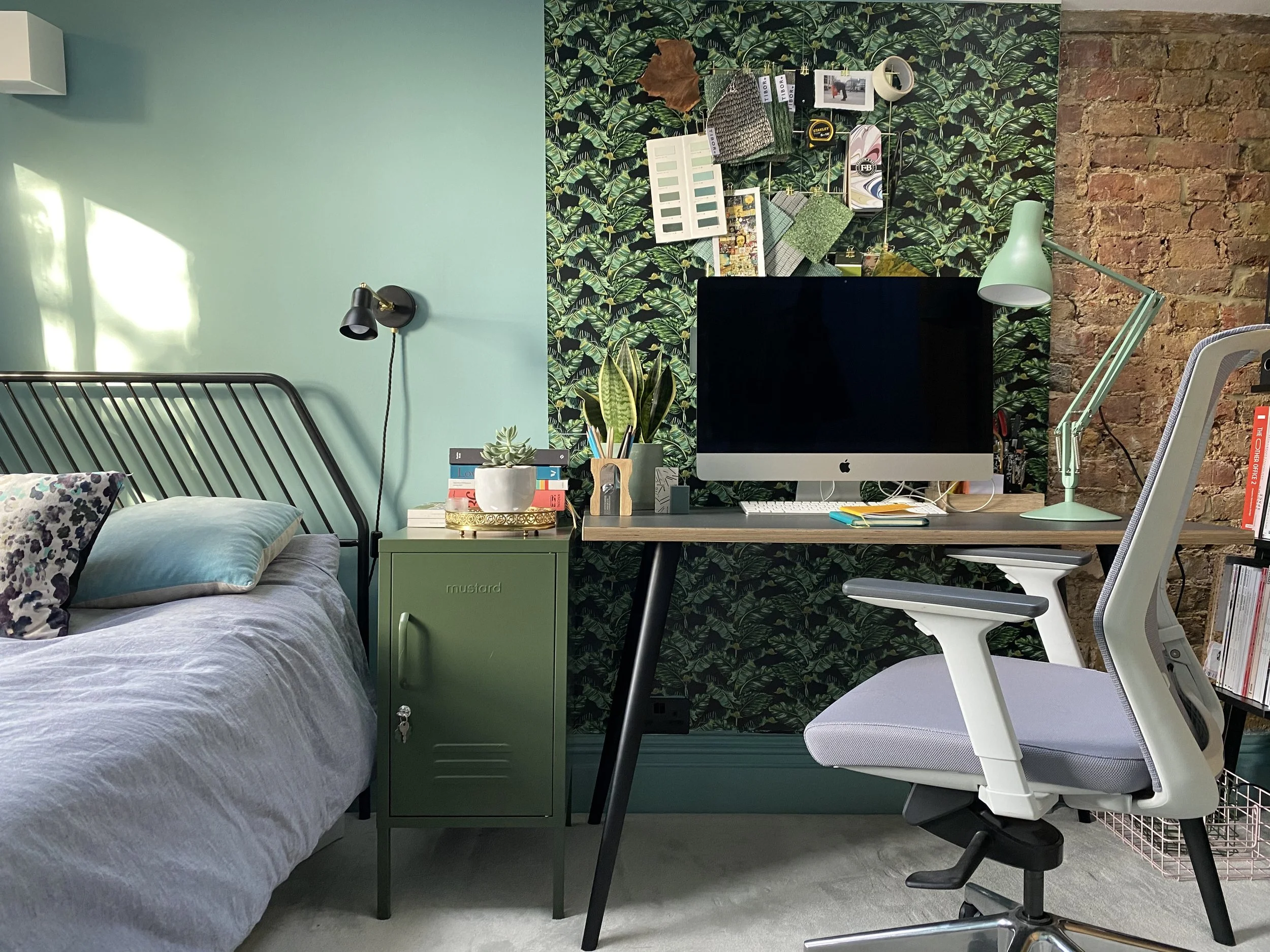 A cozy contemporary bedroom workspace with a black metal bed frame, light gray bedding with floral and solid pillows, a green nightstand with potted plants and books, a wall-mounted black reading lamp, a wooden desk with an iMac computer, a mint gree