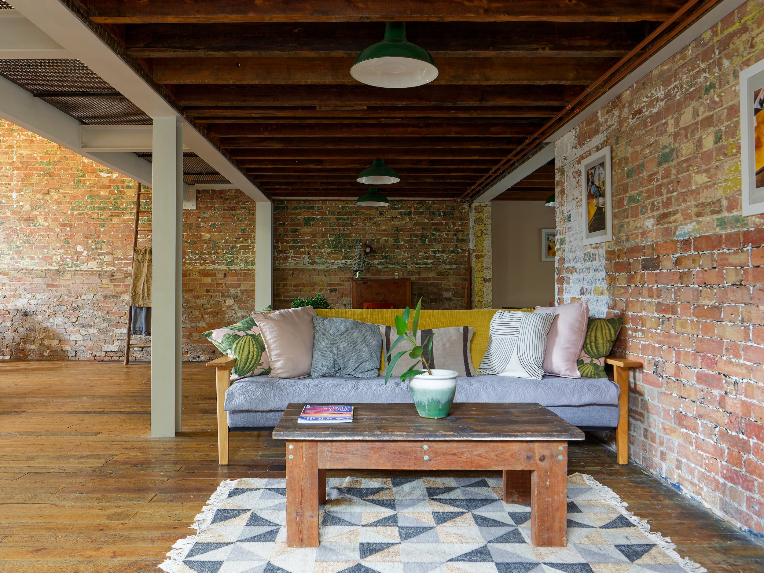 Living room with exposed brick walls, wooden floor, gray sofa with multiple pillows, rustic wooden coffee table with a potted plant and magazines, geometric-patterned rug, and framed artwork on the walls
