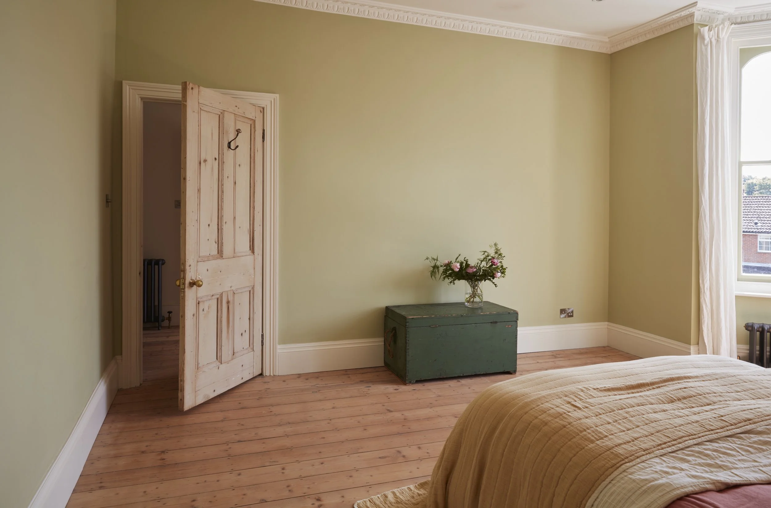 A bedroom with pale yellow walls, a wooden door partially open, a green storage chest with a glass vase of pink and white flowers on top, a window with white curtains, and a beige bedspread.