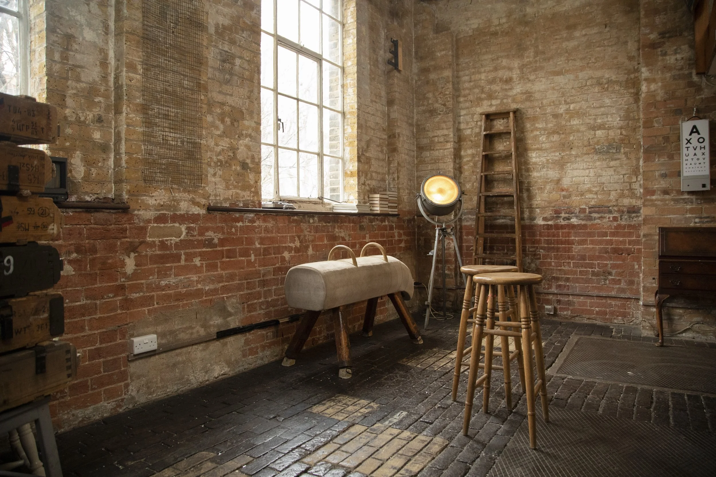 An interior room with exposed brick walls, a large window, stools, a spotlight, a ladder, and an eye chart on the wall.