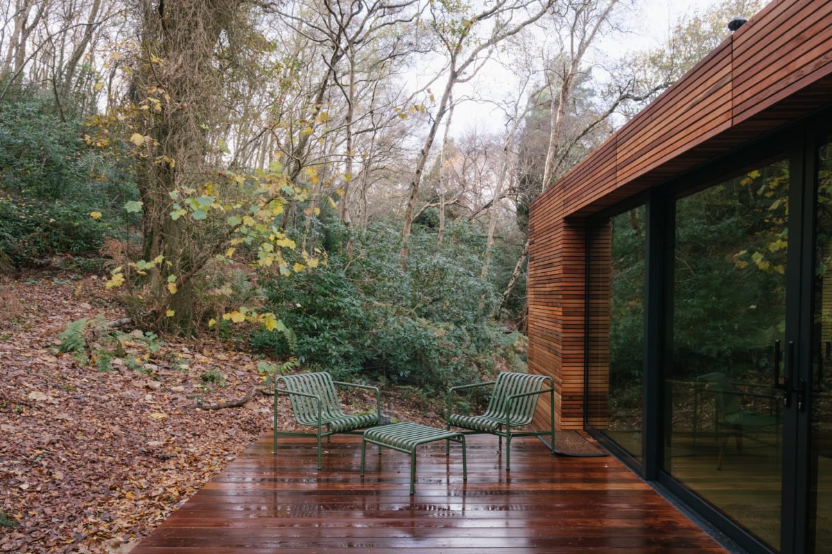 A wooden deck with wet surface and two chairs with striped cushions, located next to a house with large glass doors, surrounded by trees and foliage.