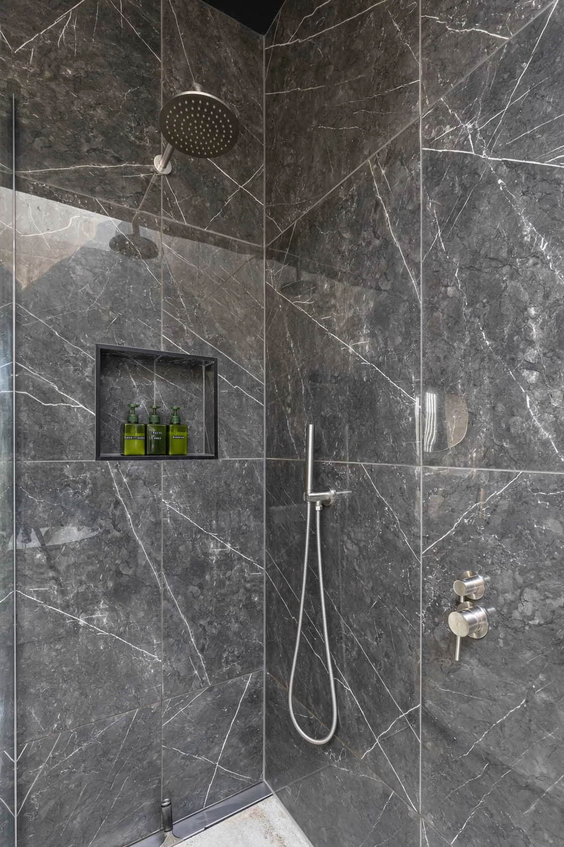 A modern shower with dark gray marble walls, a rainfall showerhead, a handheld shower wand, a built-in shampoo niche with three green bottles, and control knobs.