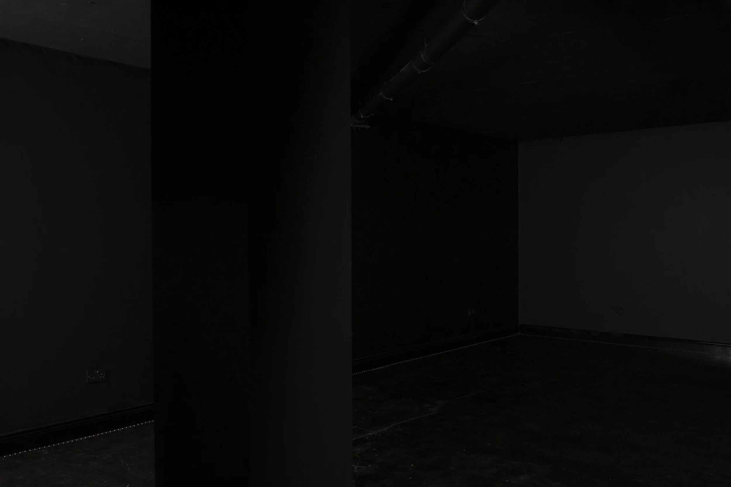 Dark, empty room with black walls, ceiling, and floor, with a black pillar in the center and a pipe running along the ceiling.