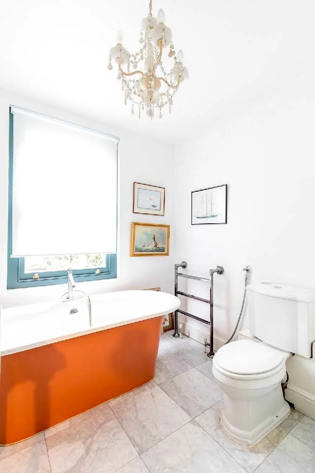 Bathroom with white walls, a bathtub with an orange exterior, a window with a white shade, framed artwork on the wall, a white toilet, and a towel rack.