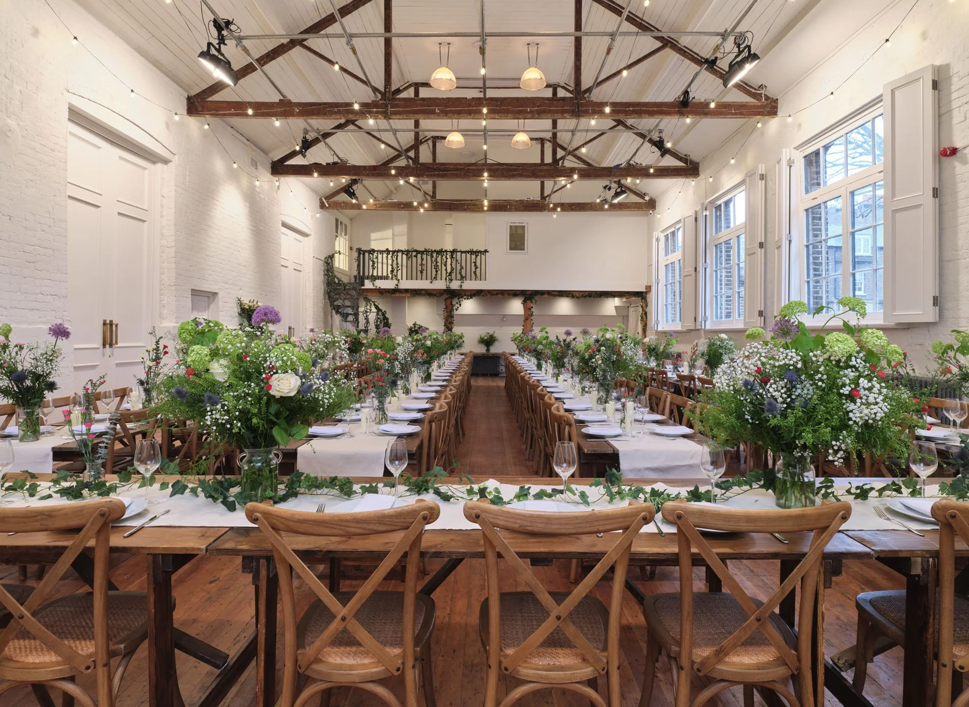 Long banquet tables with white tablecloths and wooden chairs, decorated with large floral arrangements and greenery, set for a celebration in a bright, rustic event space with exposed ceiling beams and large windows.
