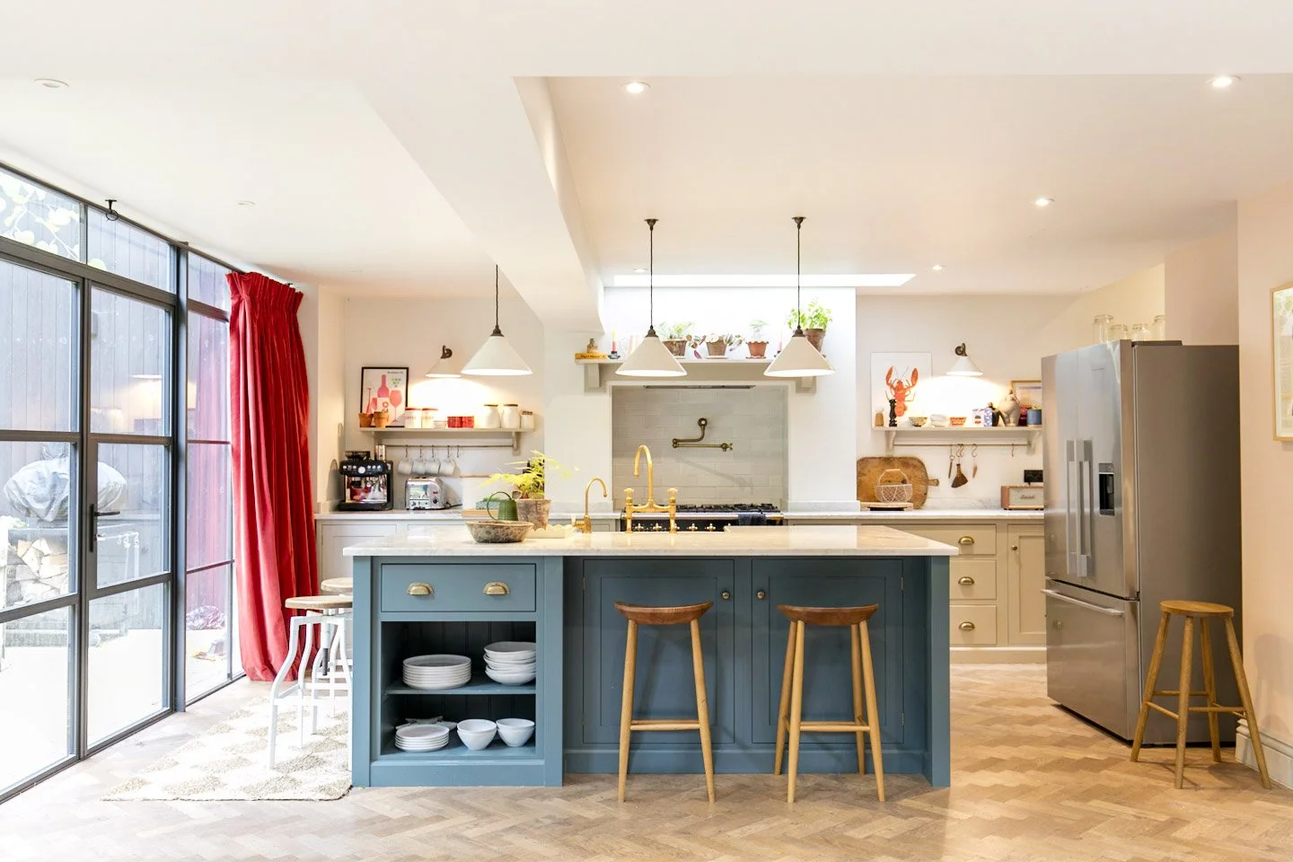 Bright kitchen with blue island, wooden stools, stainless steel refrigerator, white countertops, and red curtains.