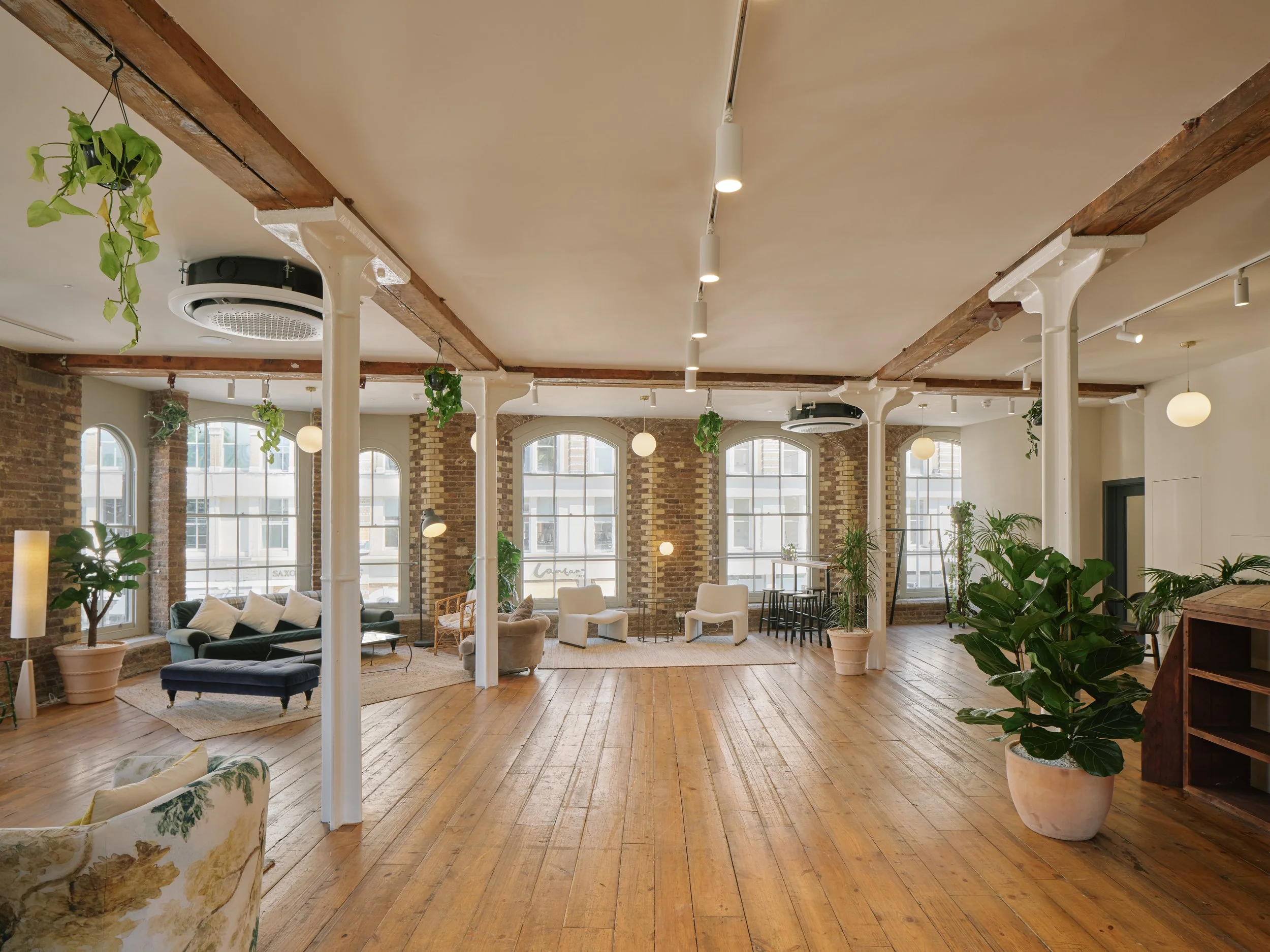 Spacious, well-lit loft with exposed brick walls, large windows, hardwood floors, and modern furniture including sofas, chairs, and potted plants.