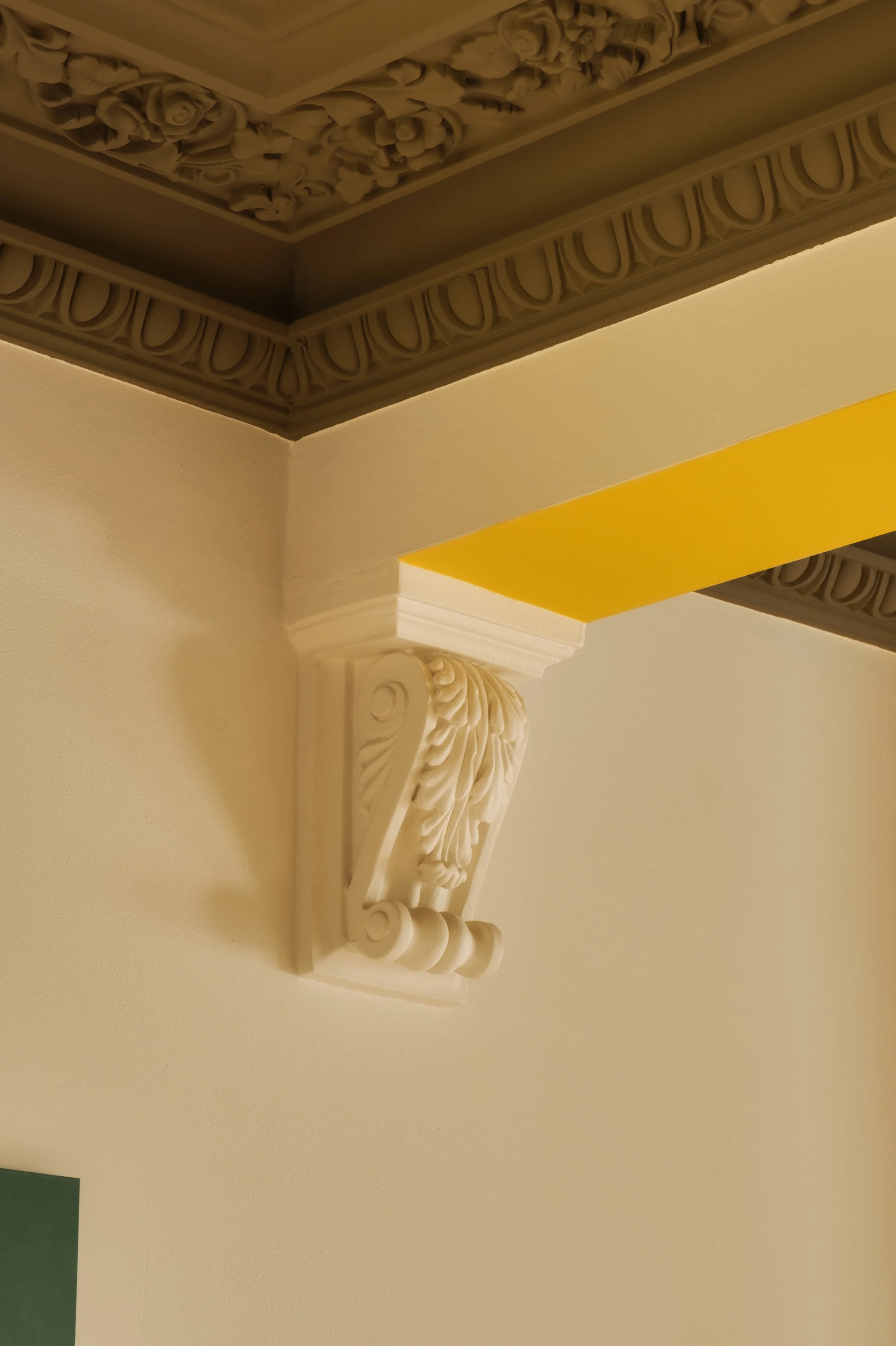 Close-up of an ornate white decorative corbel supporting a yellow-painted beam, with detailed floral and leaf patterns, beneath a ceiling with intricate molding.