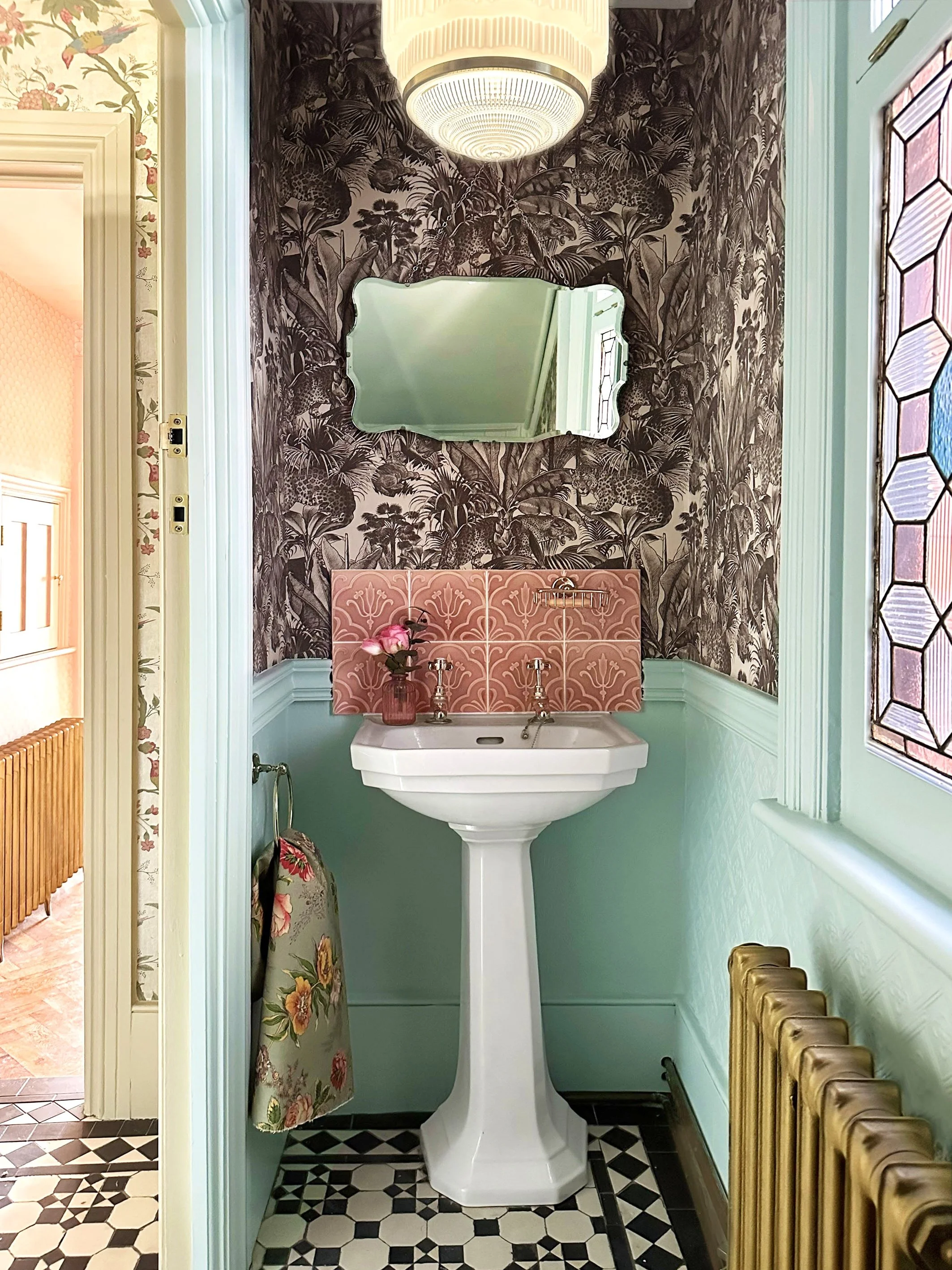 A vintage-style bathroom with a white pedestal sink, floral wallpaper, a mirror, and a stained-glass window.