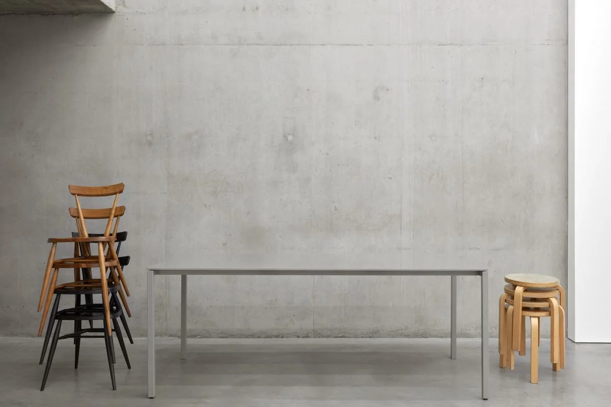 Stacked chairs on the left, a long gray table in the center, and a group of small wooden stools on the right against a plain concrete wall.