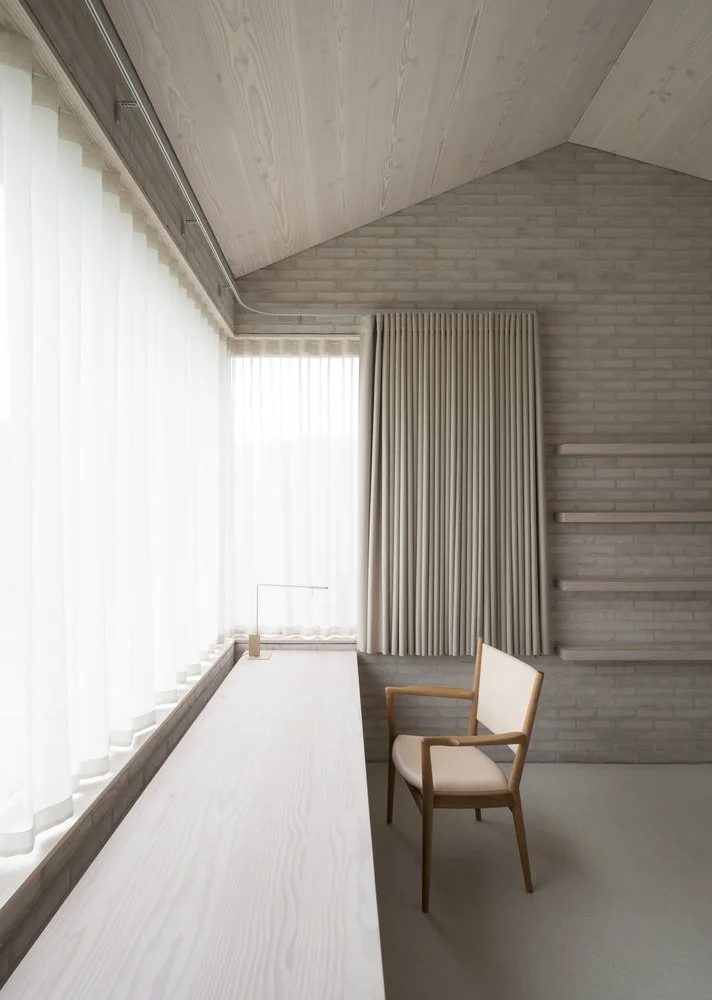 A minimalist office space with a long white desk, a beige chair, sheer white curtains, a brick wall, and shelves.