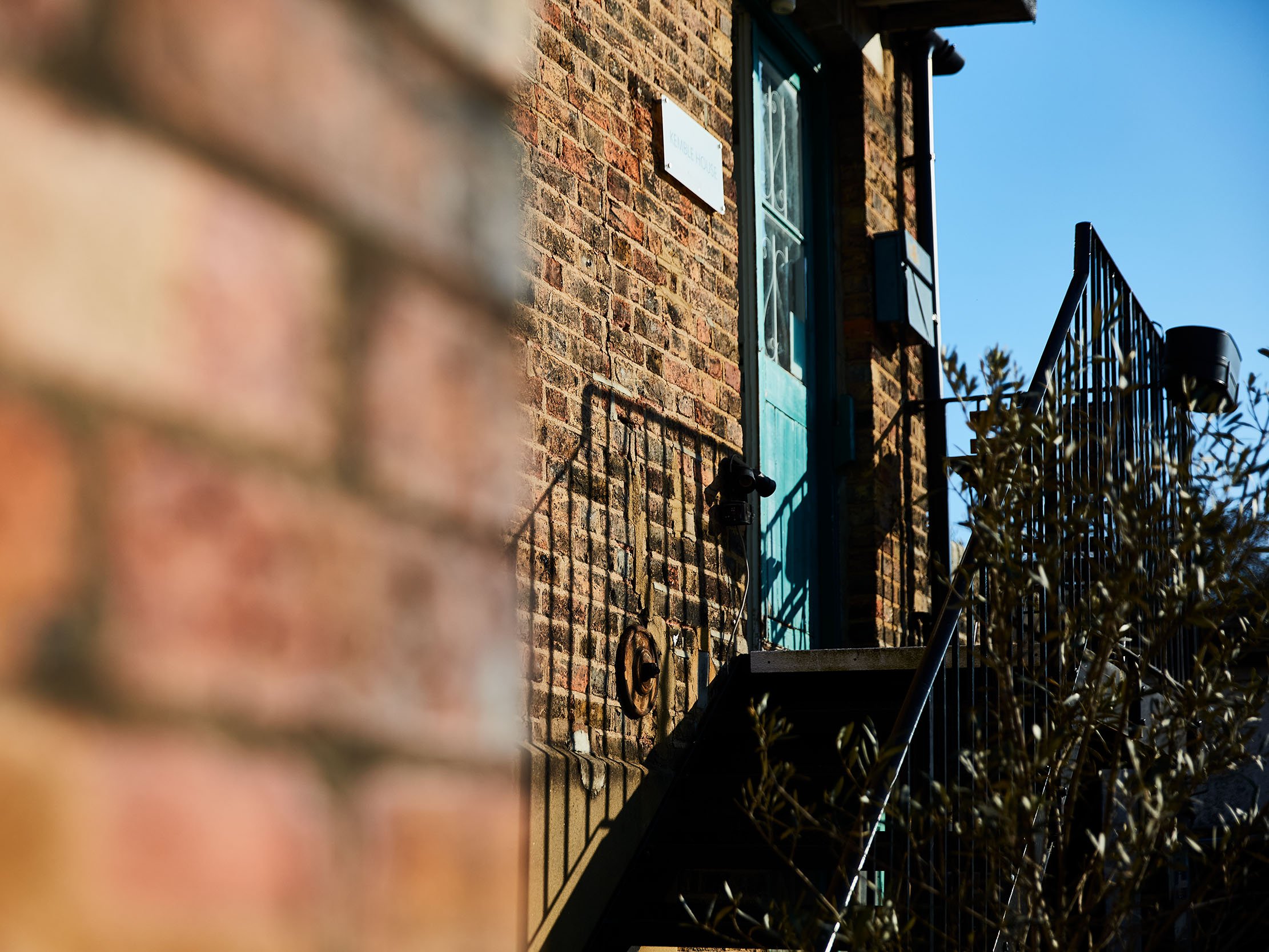 Brick building with a turquoise door, fire escape stairs, and a white sign, taken in bright daylight with a clear blue sky.
